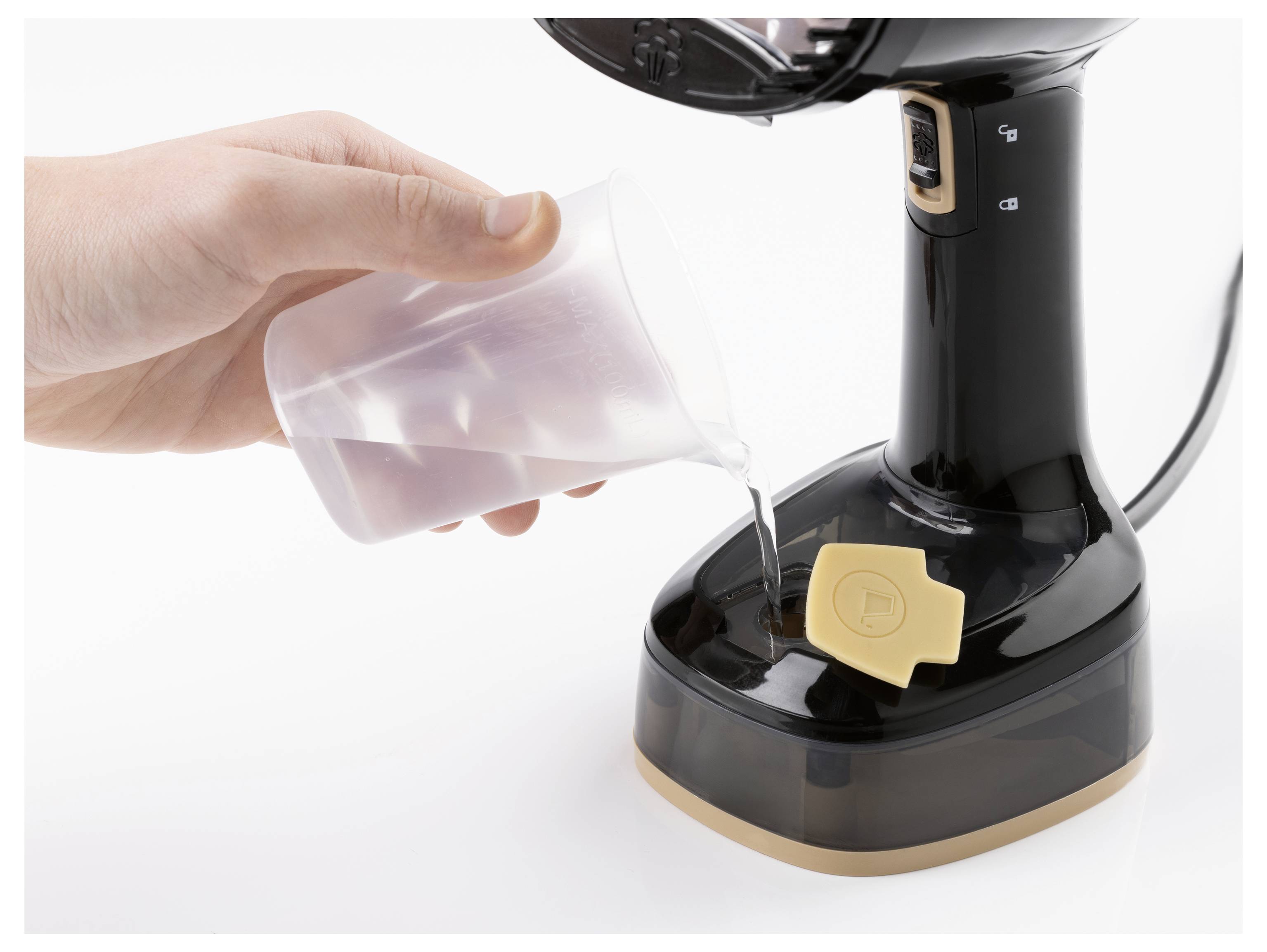 Egy kéz mérőpohárból vizet önt a fekete gőzös készülék tartályába, amely bézs gombbal rendelkezik, valószínűleg egy ruhagőzölő.