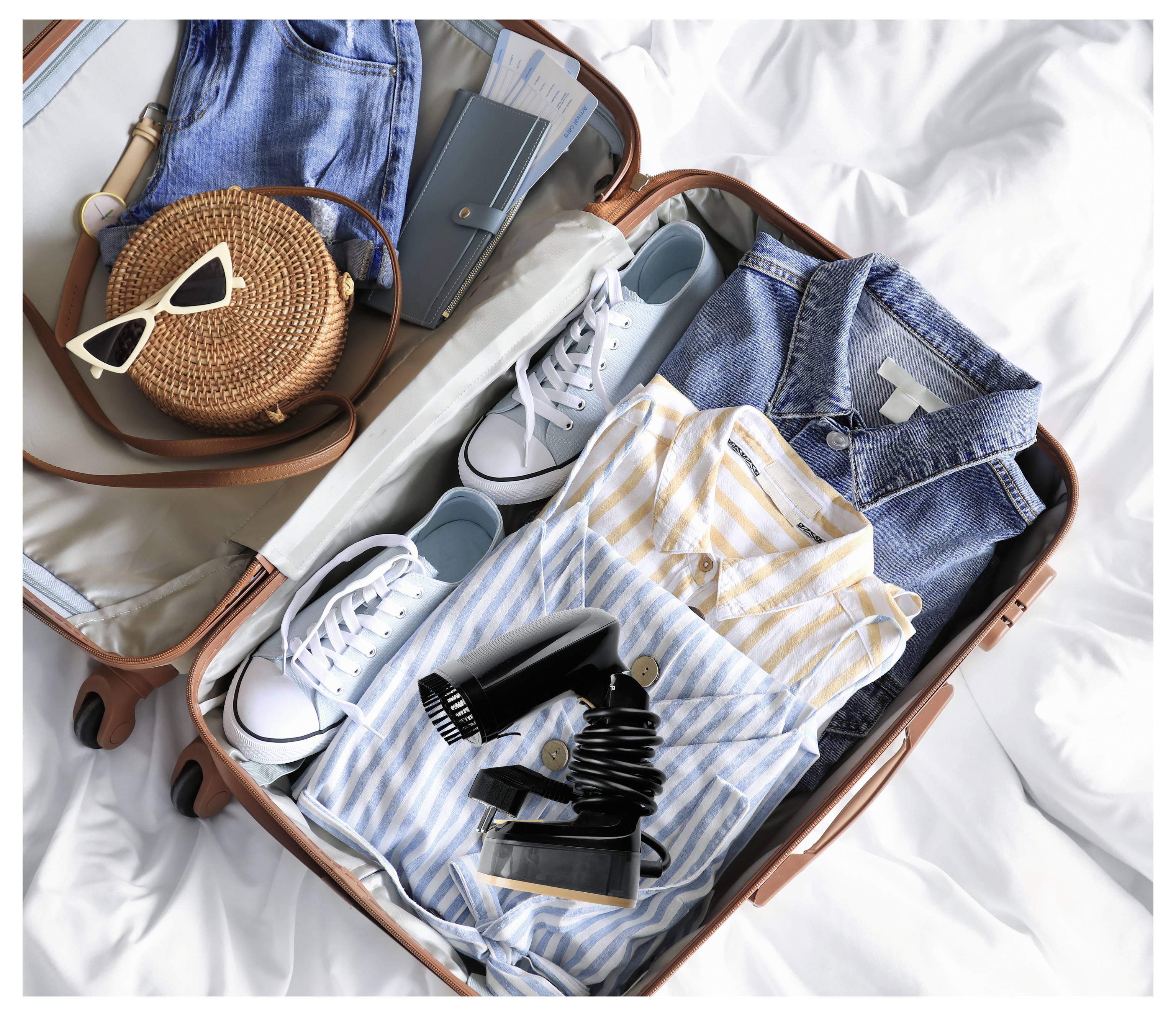 Nyitott bőrönd az ágyban rendezetten pakolva: ruhák, tornacipő, farmernadrág ing, csíkos felsőrészek, hajszárító, kerek szövött táska és napszemüveg.