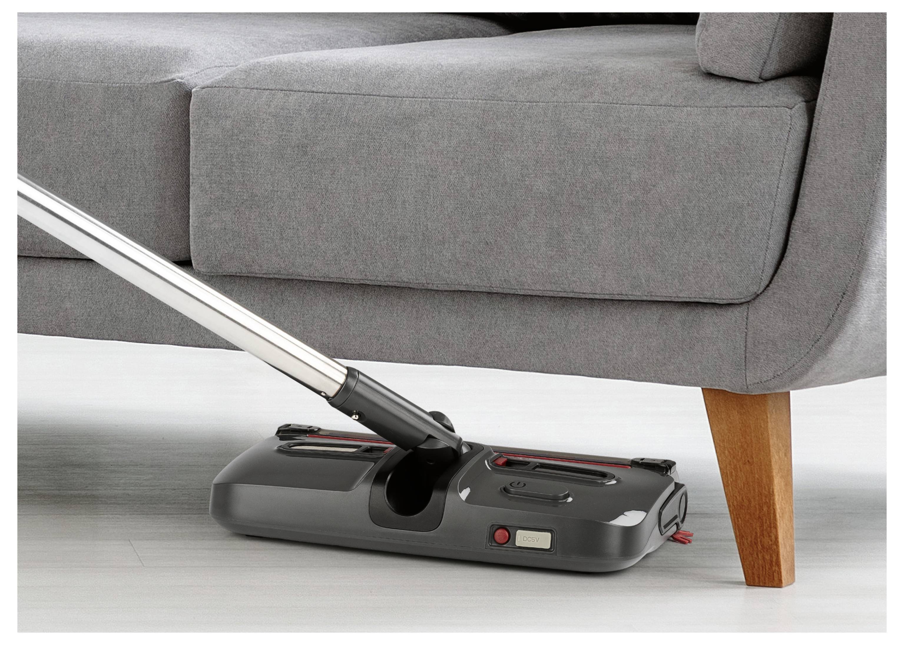 Egy hosszú nyéllel rendelkező porszívó egy szürke kanapé alatt nyúl be, amelynek fa lábai vannak. A figyelem a szoros helyeknek a tisztítására összpontosul.