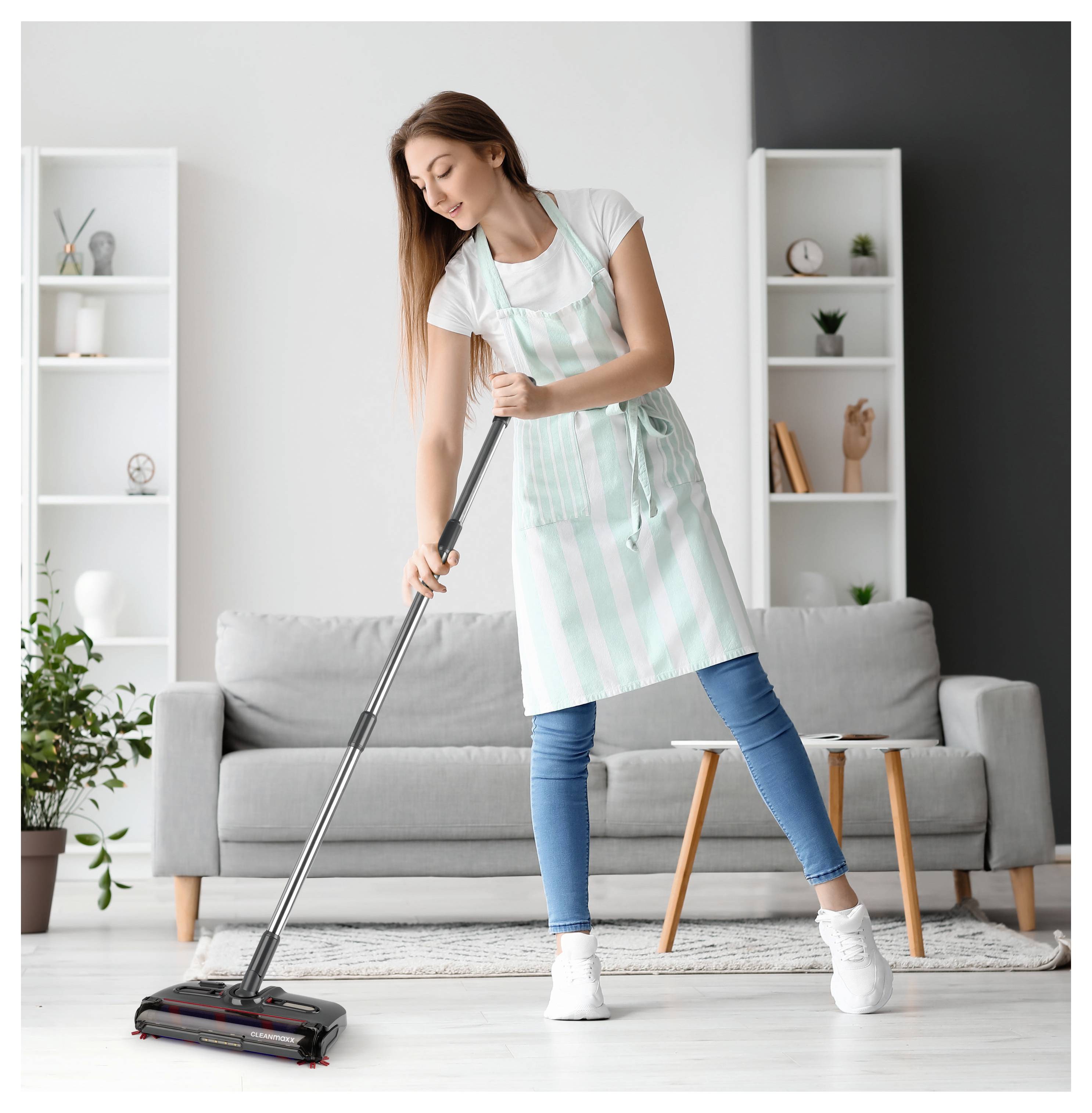 Egy csíkos kötényes nő egy modern dekorációjú nappalibolt porszívózik, amely szürke kanapét, növényeket és egy könyvespolcot tartalmaz.