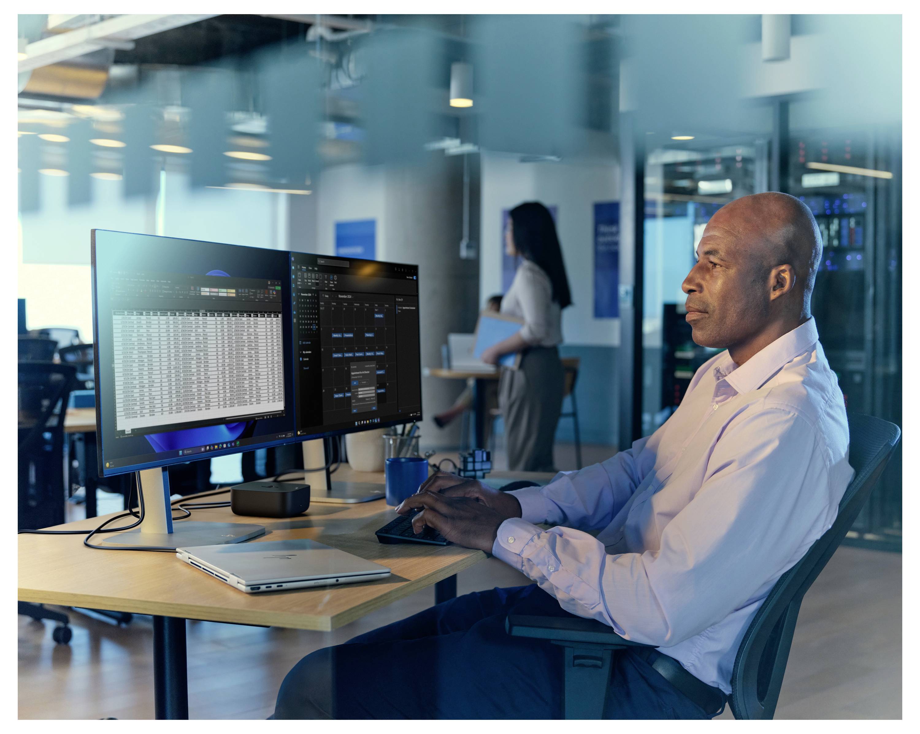 Egy személy videóhívásban több résztvevővel, akik egy számítógép-monitor kijelzőjén jelennek meg, miközben egy másik képernyőn táblázatkezelő adatok láthatók.
