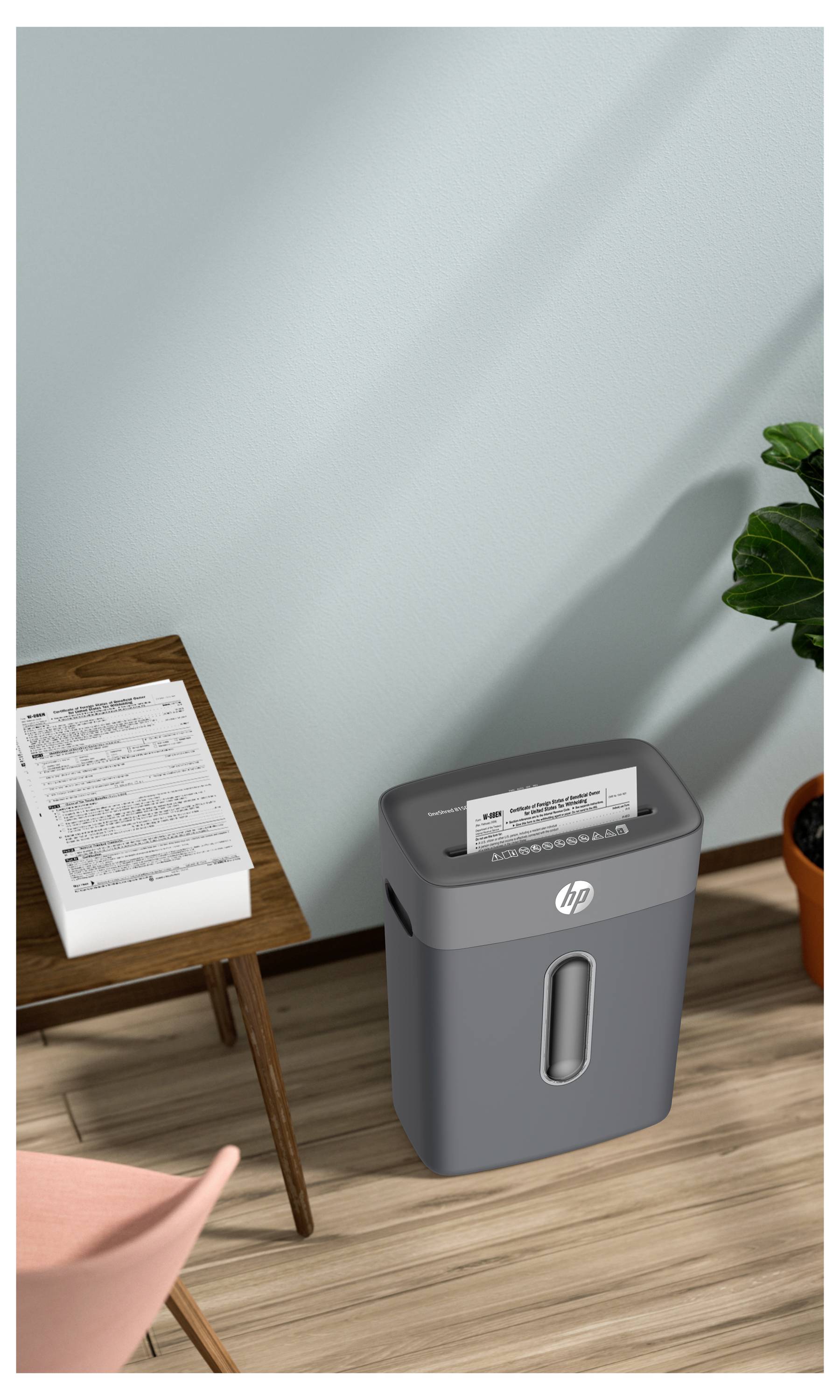 Egy dokumentum aprító egy papírcsomó mellett egy faasztalon. Egy cserépben lévő növény közelében, mindez egy világoskék fal előtt.