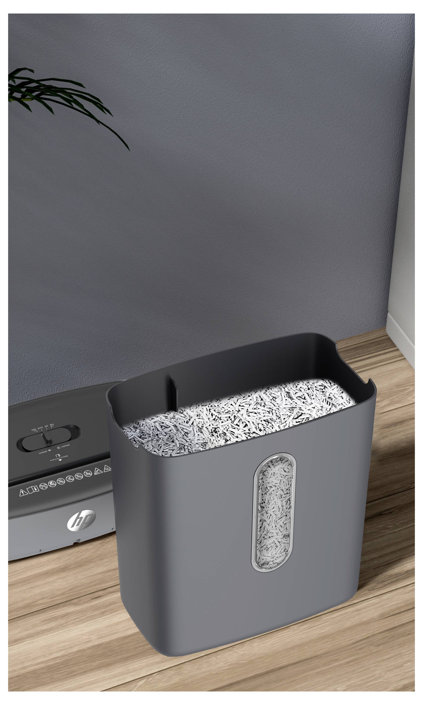 Egy szürke papírmegsemmisítő, amely megsemmisített papírral van feltöltve, fa padlón helyezkedik el egy fal mellett, közvetlenül egy kis növény közelében a bal felső sarokban.