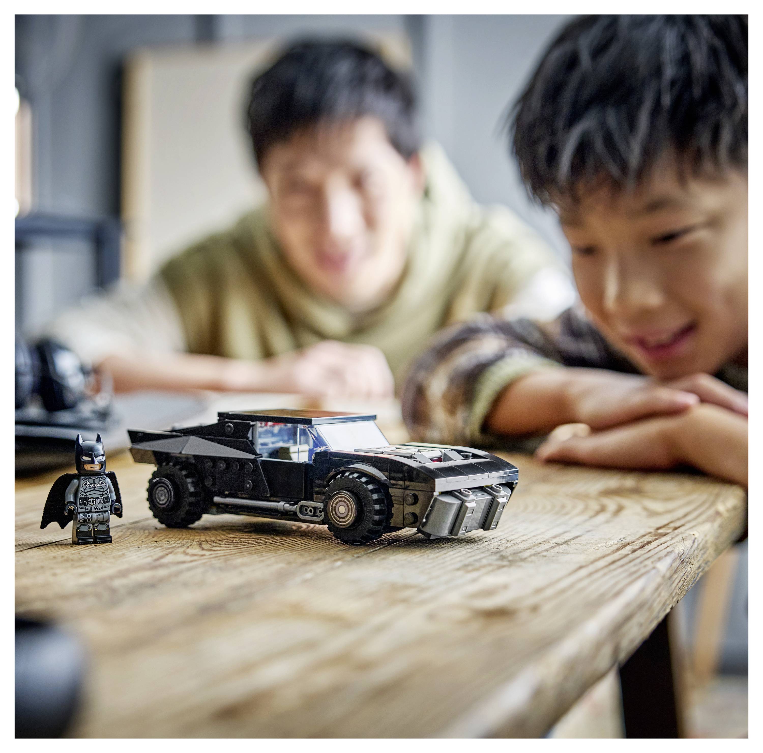 Egy mosolygó gyermek egy fazokon asztalon a Batmobiléra hasonlító játékautóra néz, az háttérben egy felnőtt boldog tekintettel figyeli.
