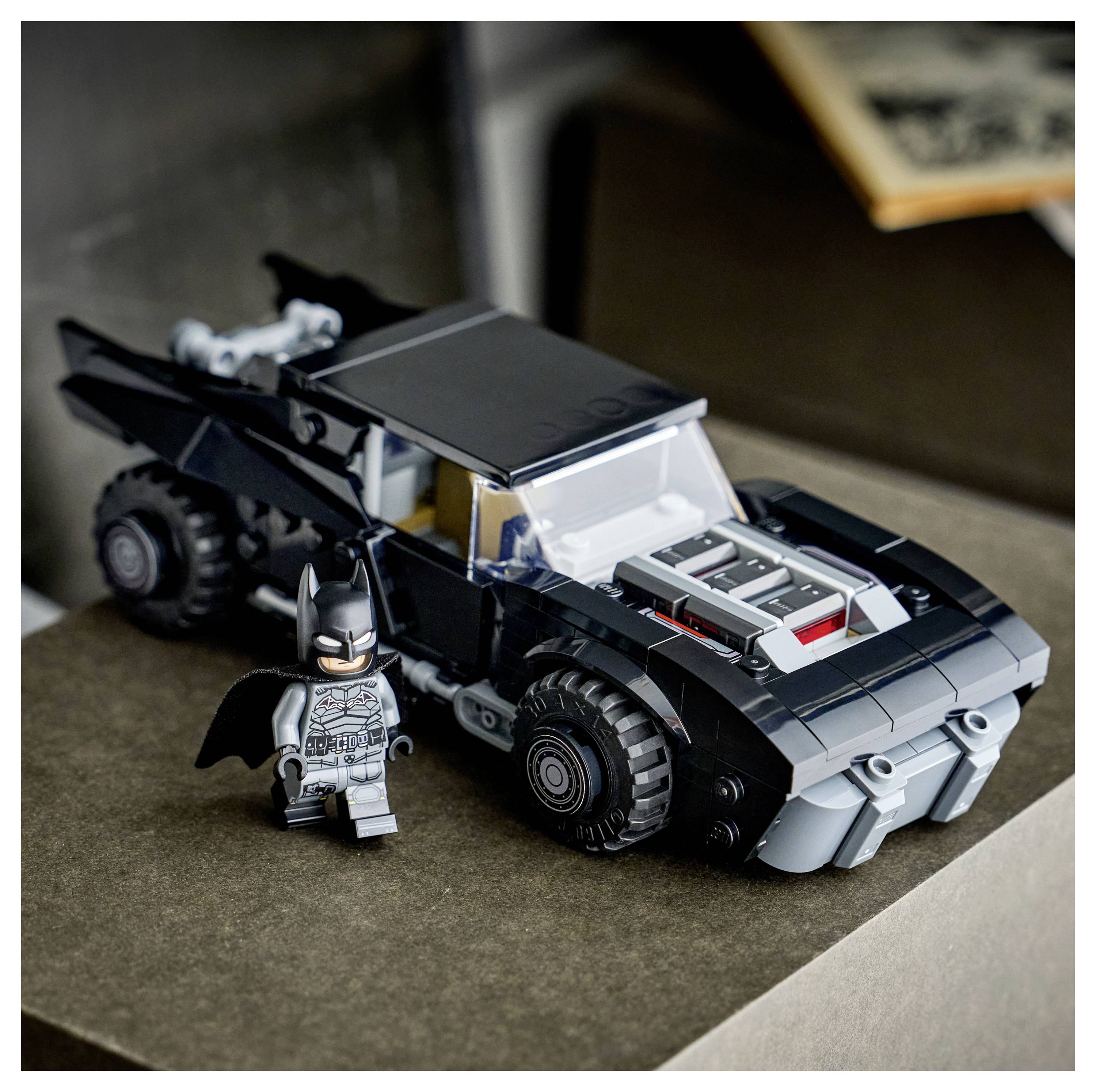 Egy fekete játékautó ezüst részletekkel egy felületen áll, mellette egy kisméretű játékfigura, amely fekete szuperhősköltözékben van.