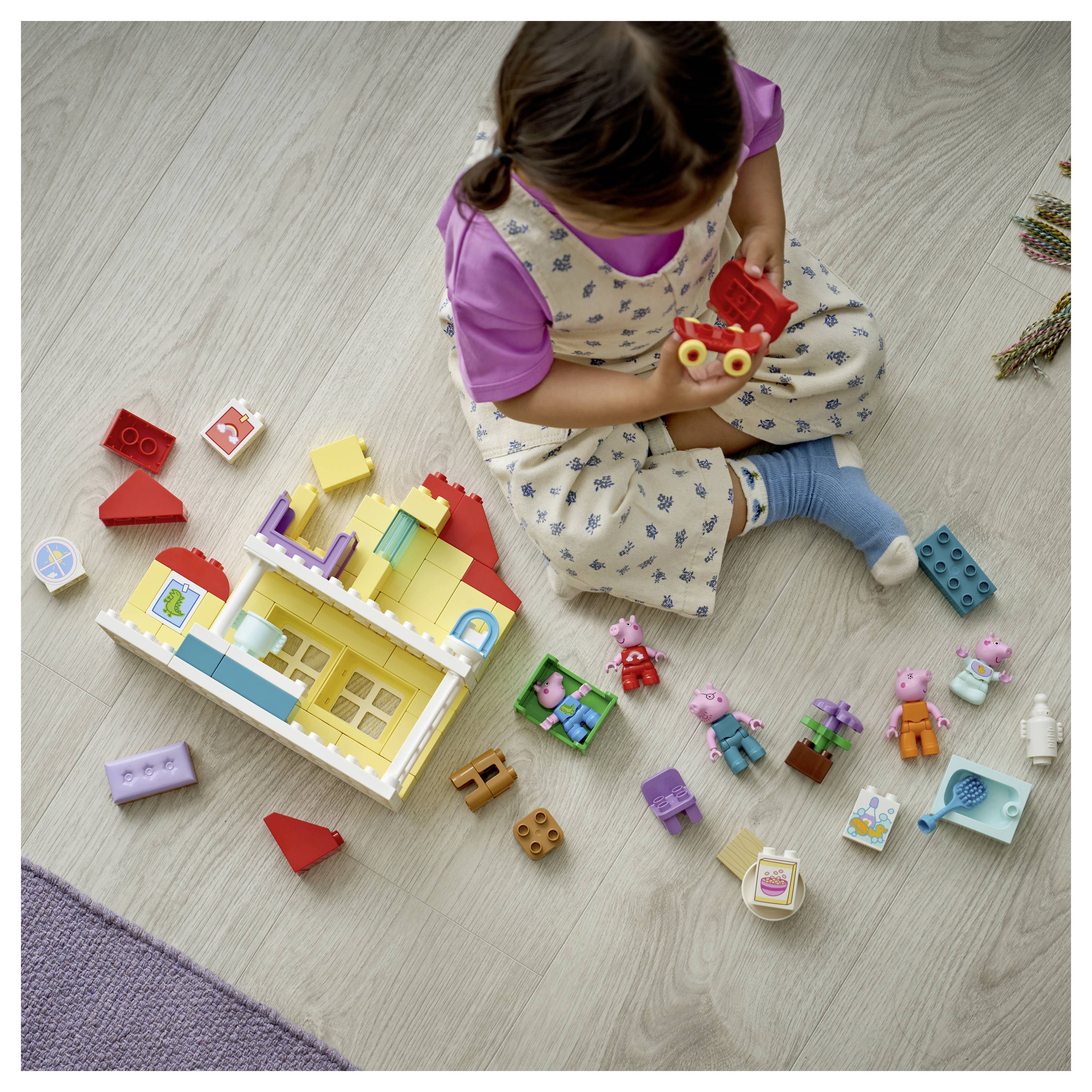 Egy gyermek a padlón ül és építőkockákkal és játékfigurákkal játszik, figyelmesen nézve a kezében tartott kis piros játékra, számos szétszórt, színes darab között.