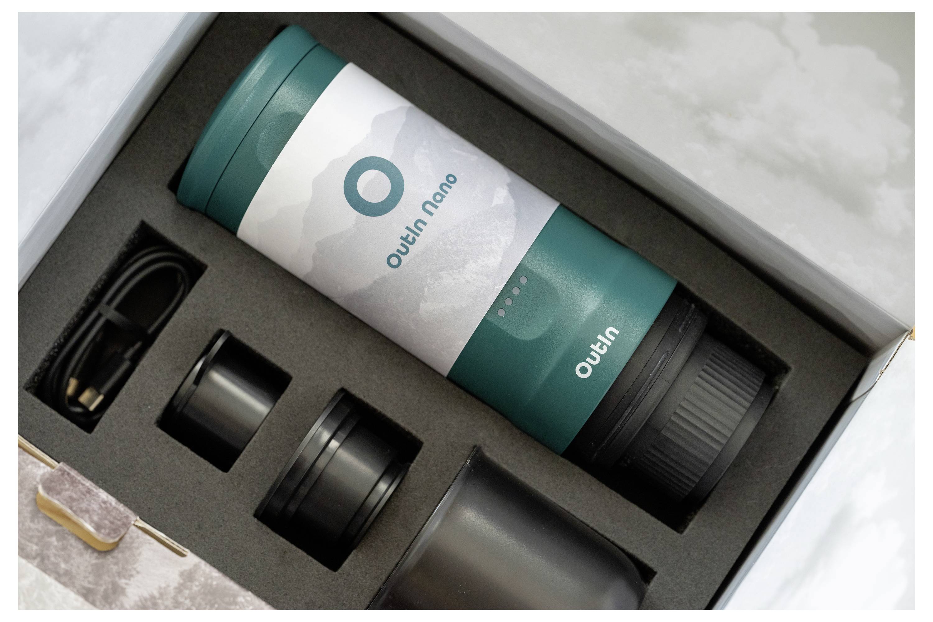 Egy tealszín és fehér hordozható vákuum termosz, amelyen az "Outin Nano" felirat látható, egy fekete dobozban, amely rekeszeket tartalmaz, beleértve egy kiegészítőt és egy sapkát.