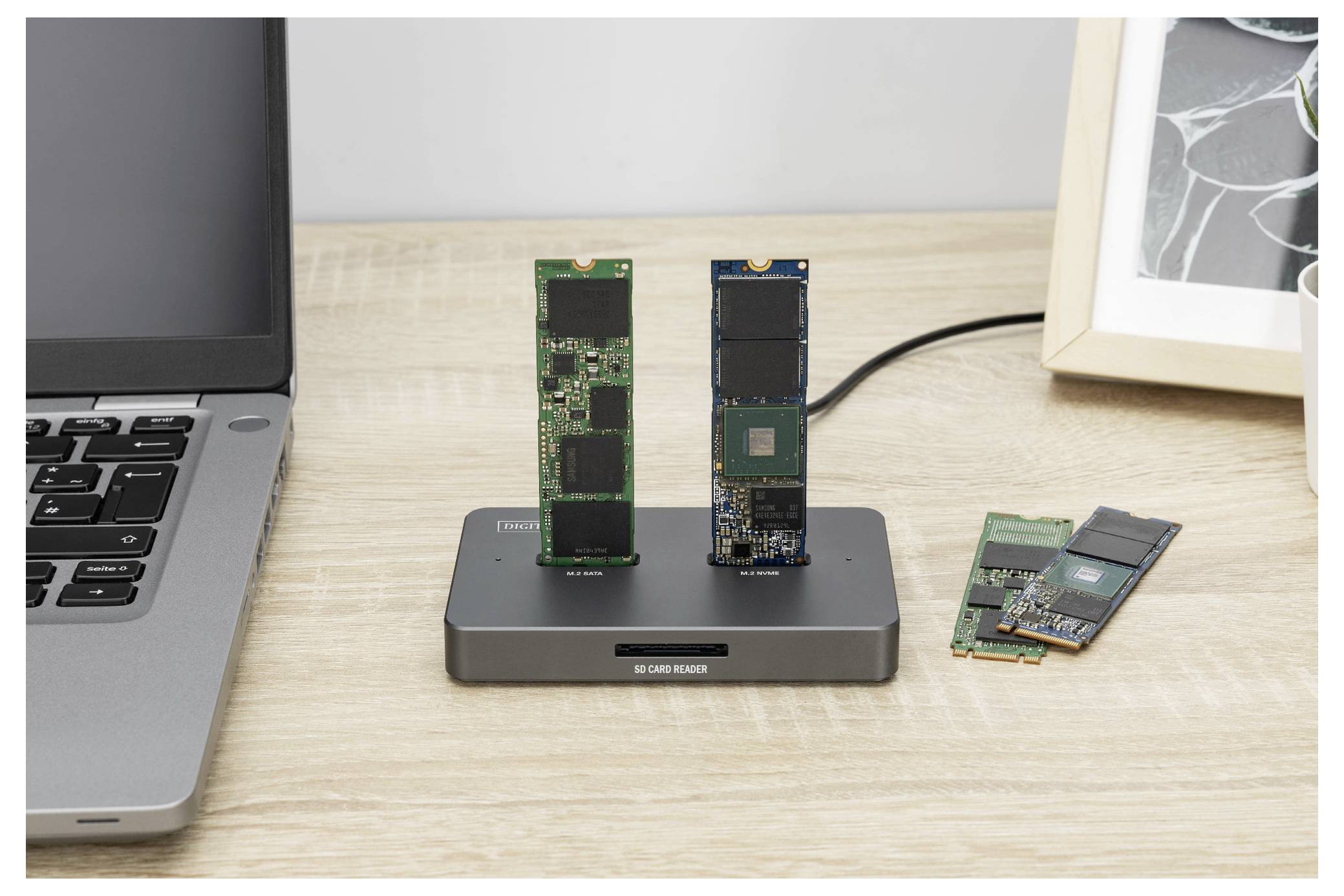 Egy dokkolóállomás két NVMe SSD-t tart függőlegesen; egy laptop a bal oldalon, és egy keretezett fénykép részben látható a jobb oldalon.