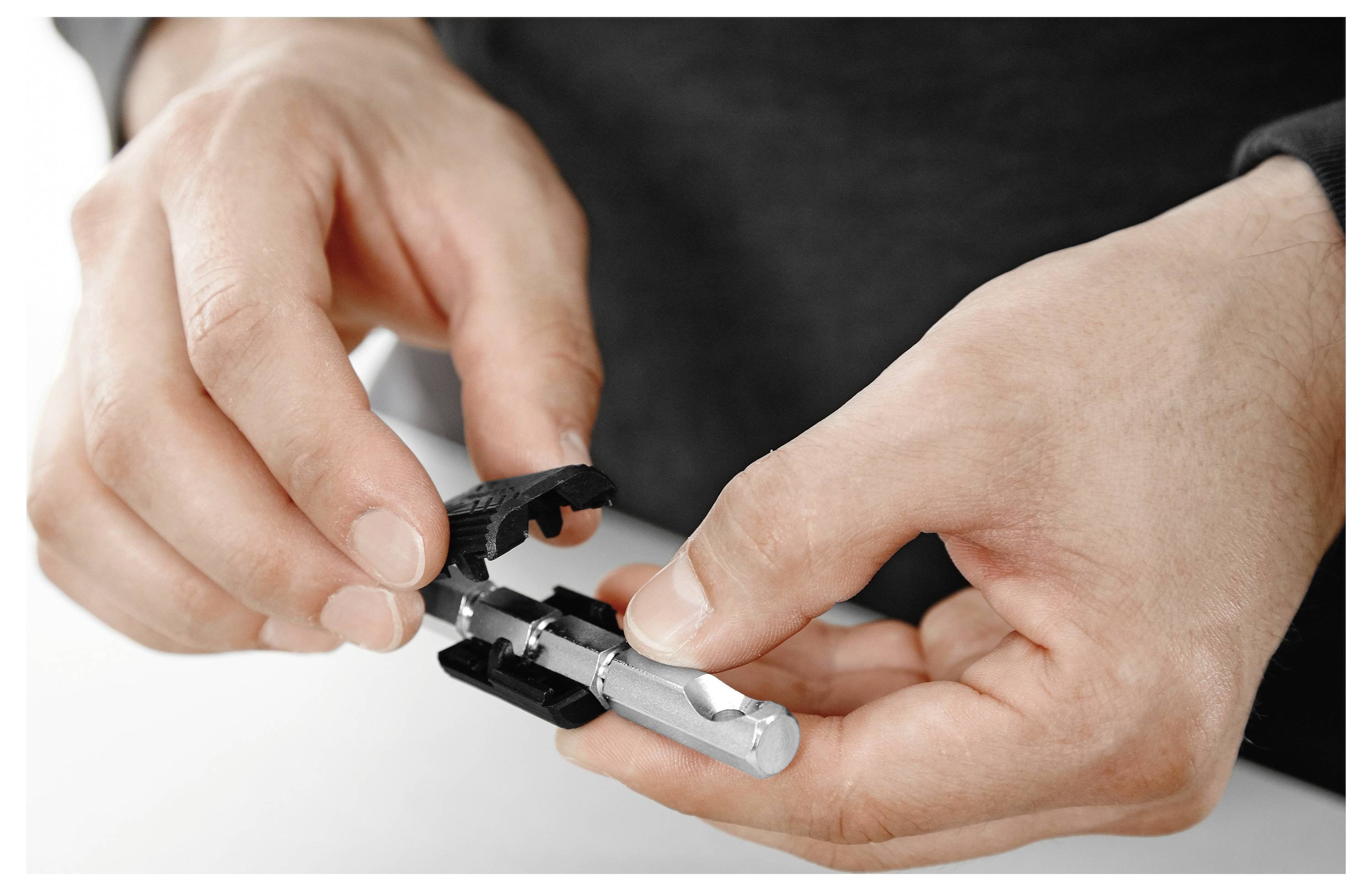Kezek egy kis fekete és ezüst eszközt, esetleg szerszámot vagy elektronikus alkatrészt szerelnek össze fehér felületen.