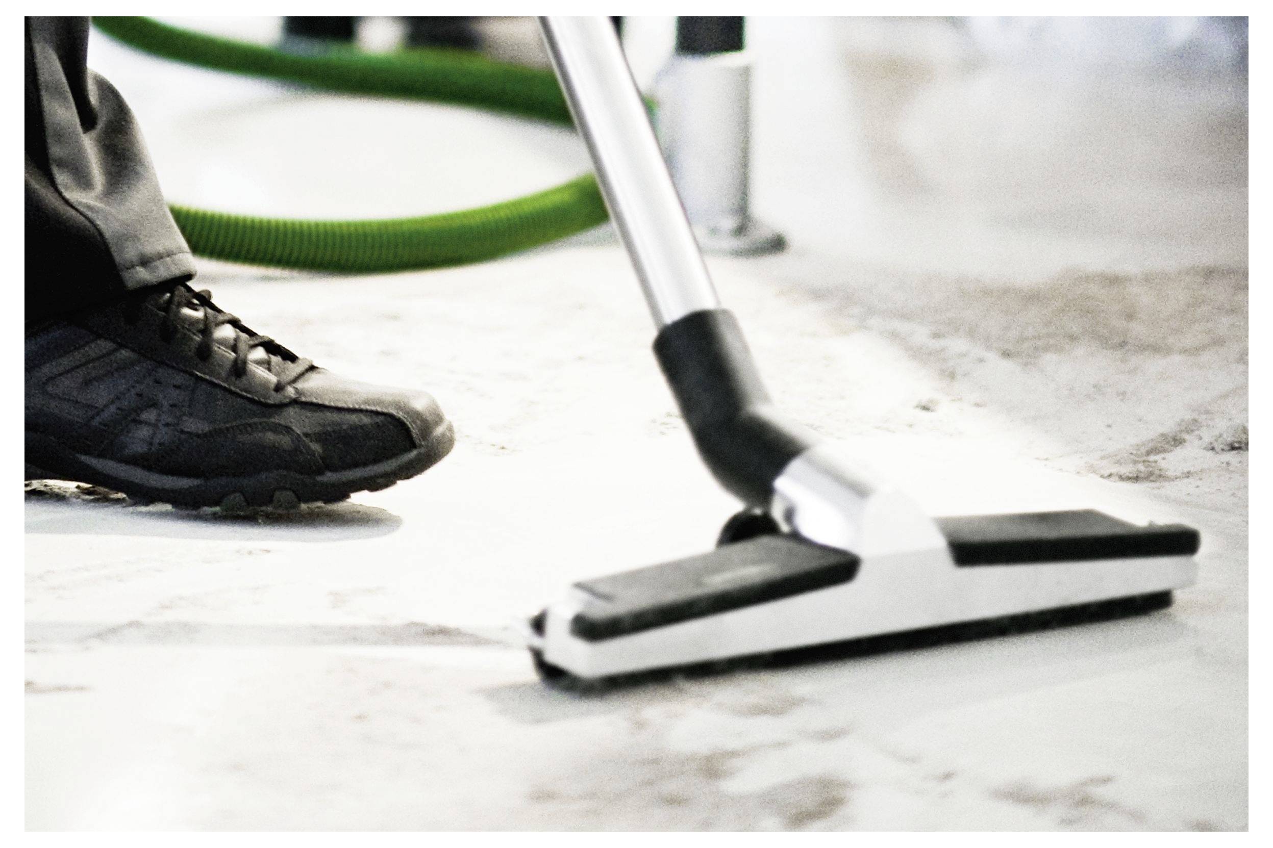 'Egy személy világos színű padlót takarít porszívóval, fekete és ezüst porszívó csővel, a szerszám és a cipő közeli képe.'