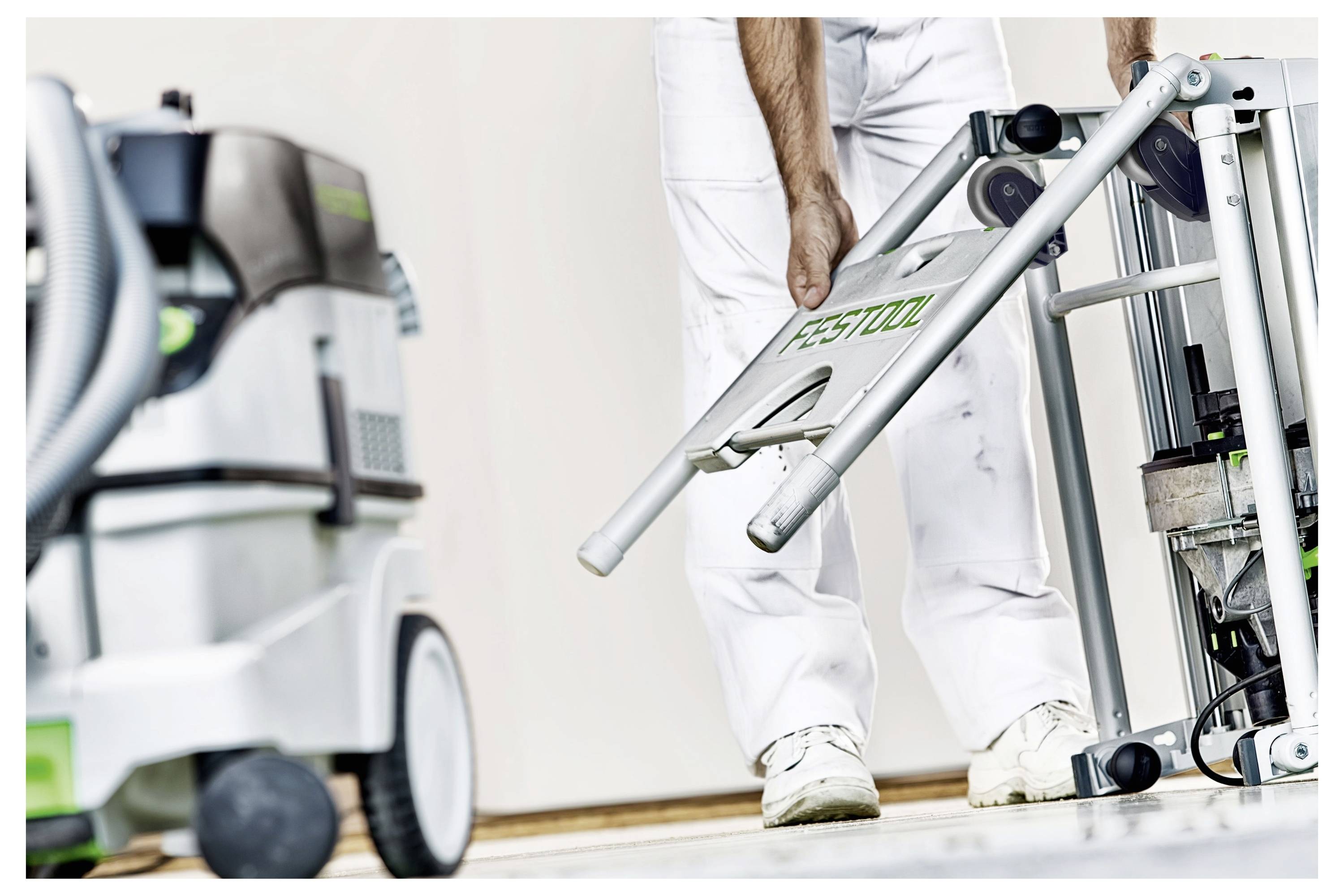 Egy fehér munkaöltözetet viselő személy egy Festool gépet szerel össze vagy állít be, az előtérben pedig egy másik Festool porszívó látható.