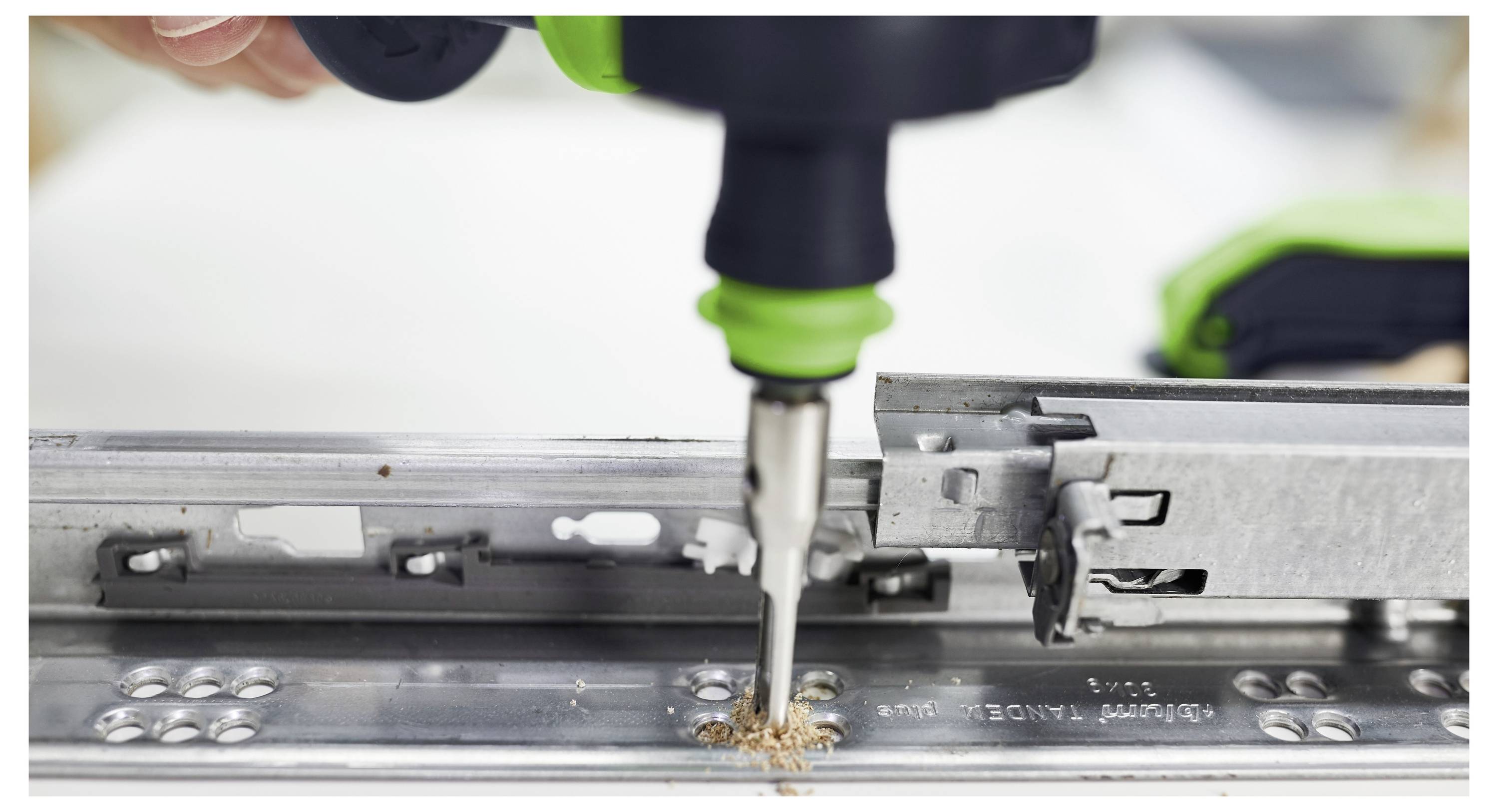 Egy fúró közelről, amely lyukat fúr egy fémvonalba, faforgács látható. A fúrót egy kéz tartja, közel a fókuszhoz.