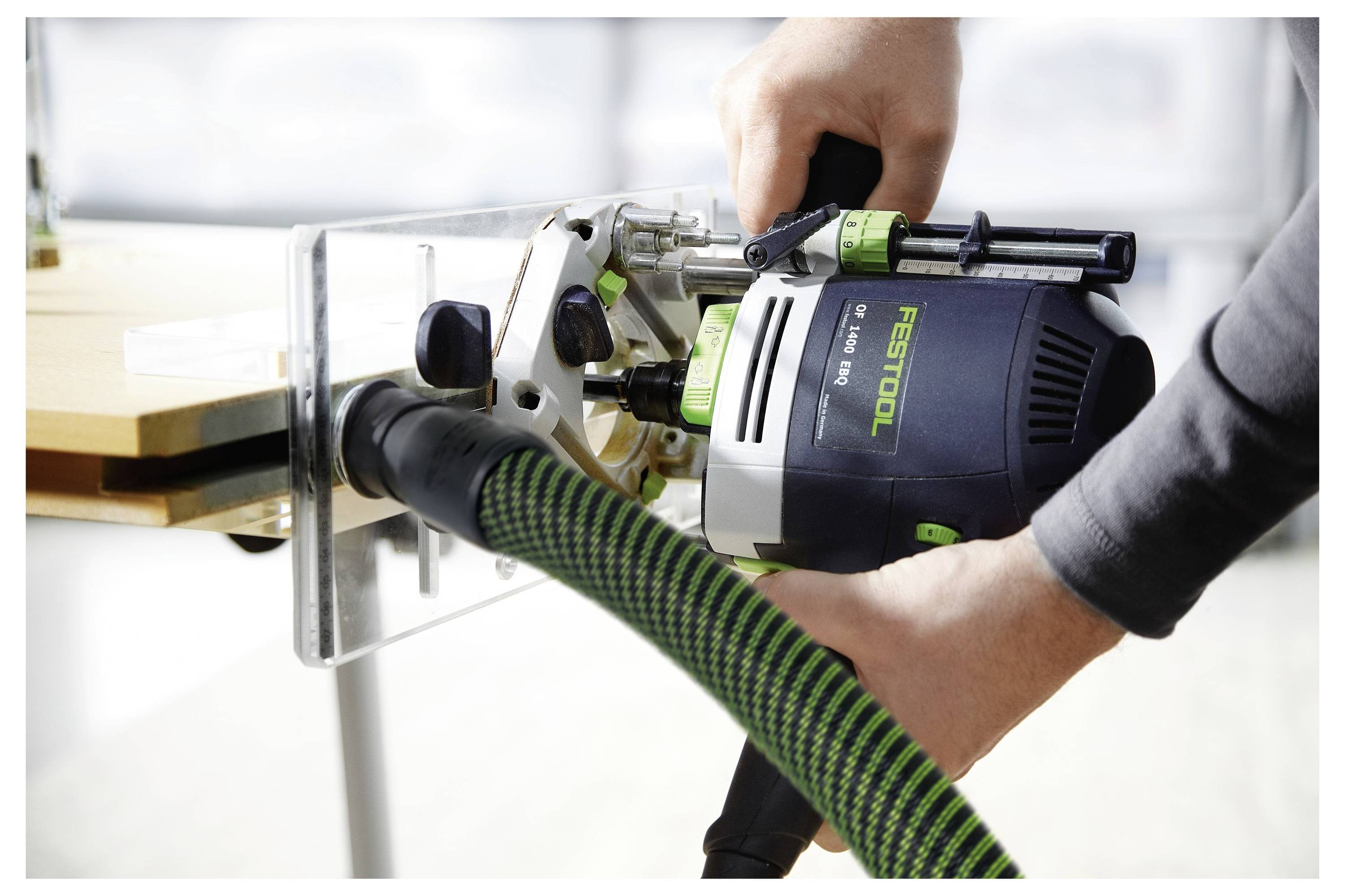 Egy személy Festool felsőmaró gépet használ egy fa felületen, és egy zöld-fekete csővel csatlakoztatja azt egy porfelvételő rendszerhez.