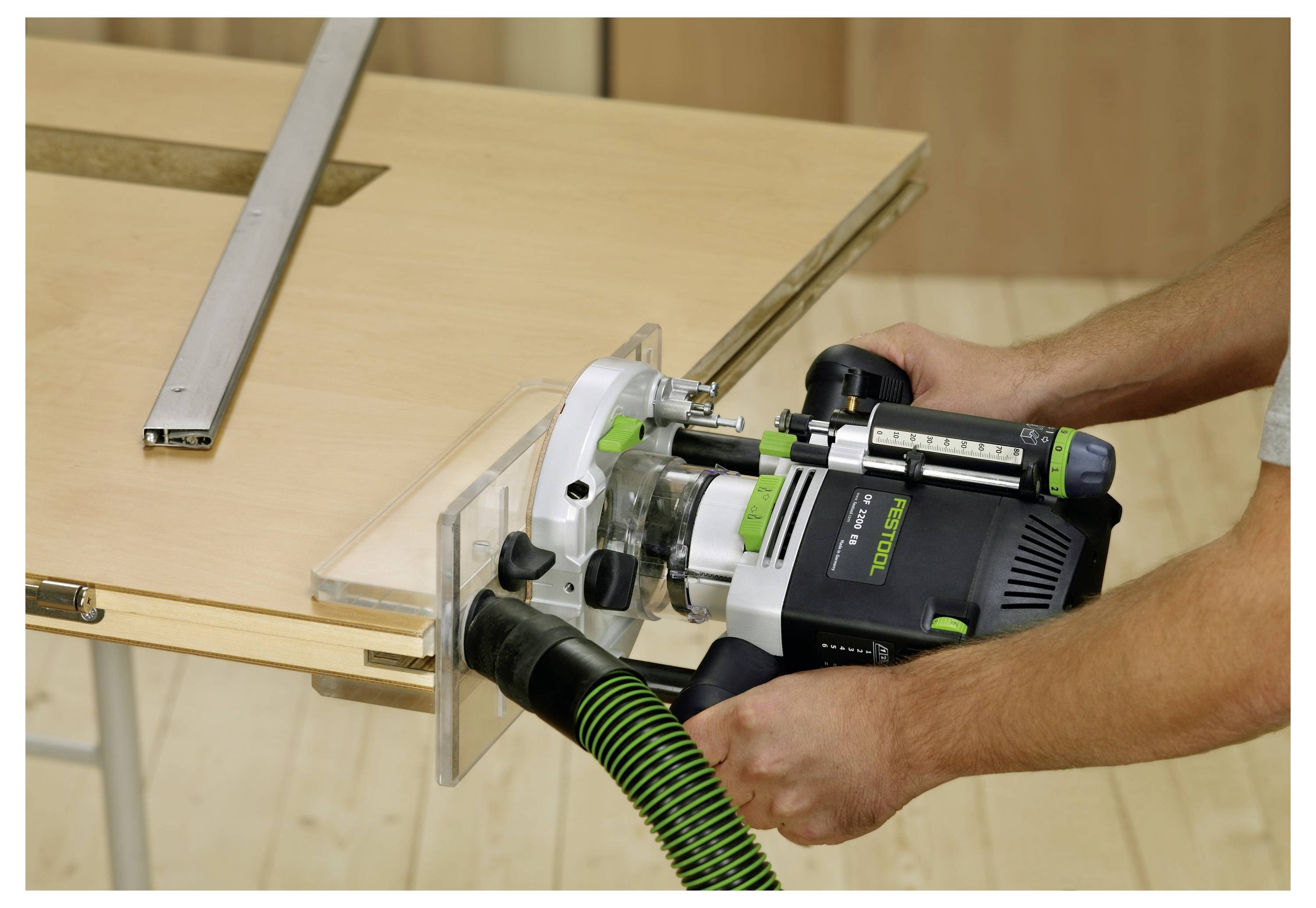 Egy személy elektromos marógépet használ egy fadeszkán, egy fém vezetősín látható az asztalon, műhelykörnyezetben.