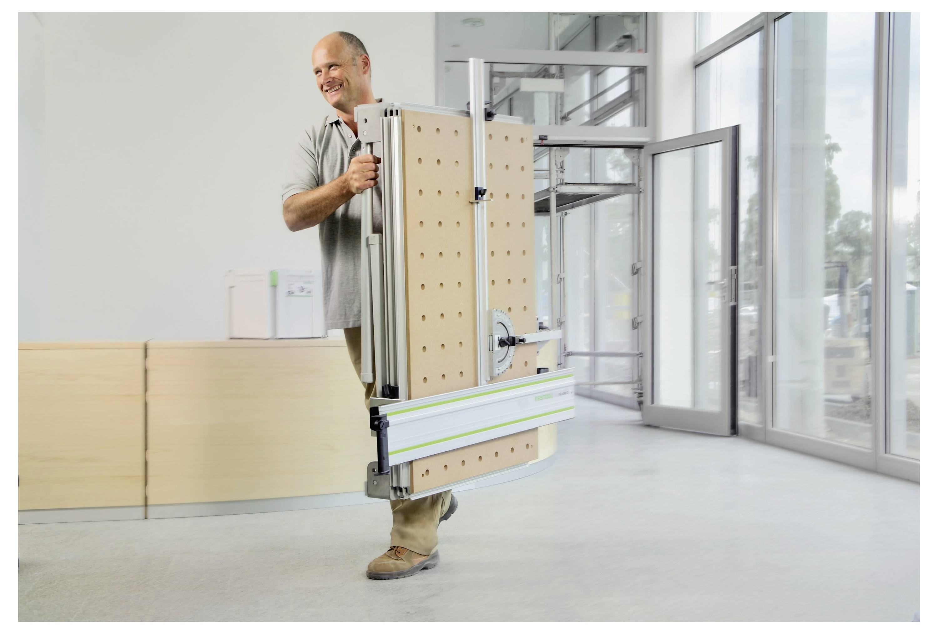 Egy mosolygó személyt ábrázoló kép, aki egy összecsukható, hordozható munkapadot hordoz pegboard oldalakkal egy világos, modern irodai térben üveges ablakokkal.