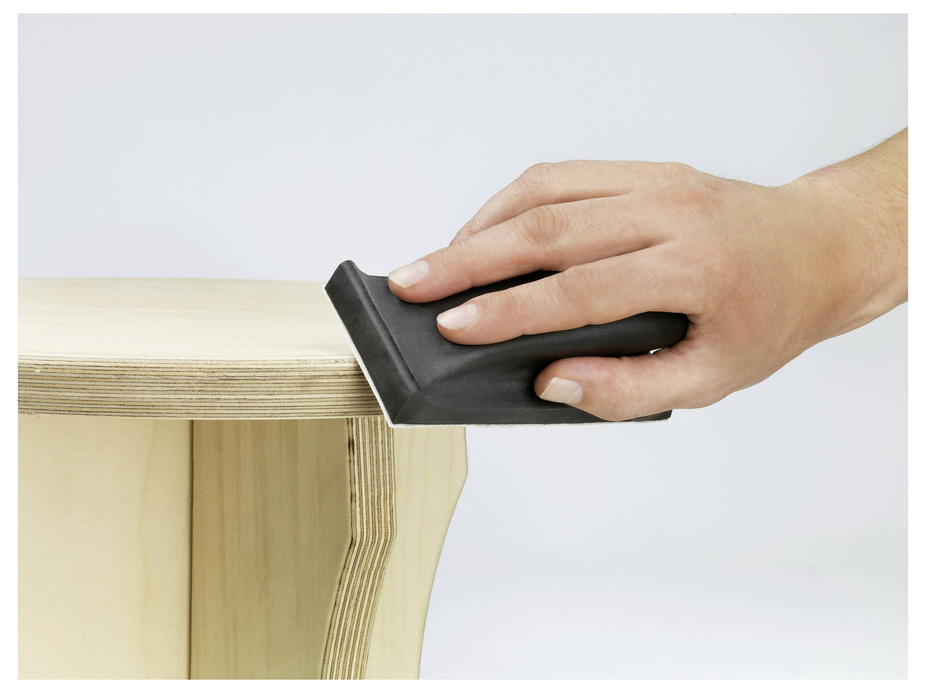 Egy kéz egy fekete csiszolóblokkkal a fa szék szélét csiszólja, simítva a felületet, egyszerű világos háttér előtt.