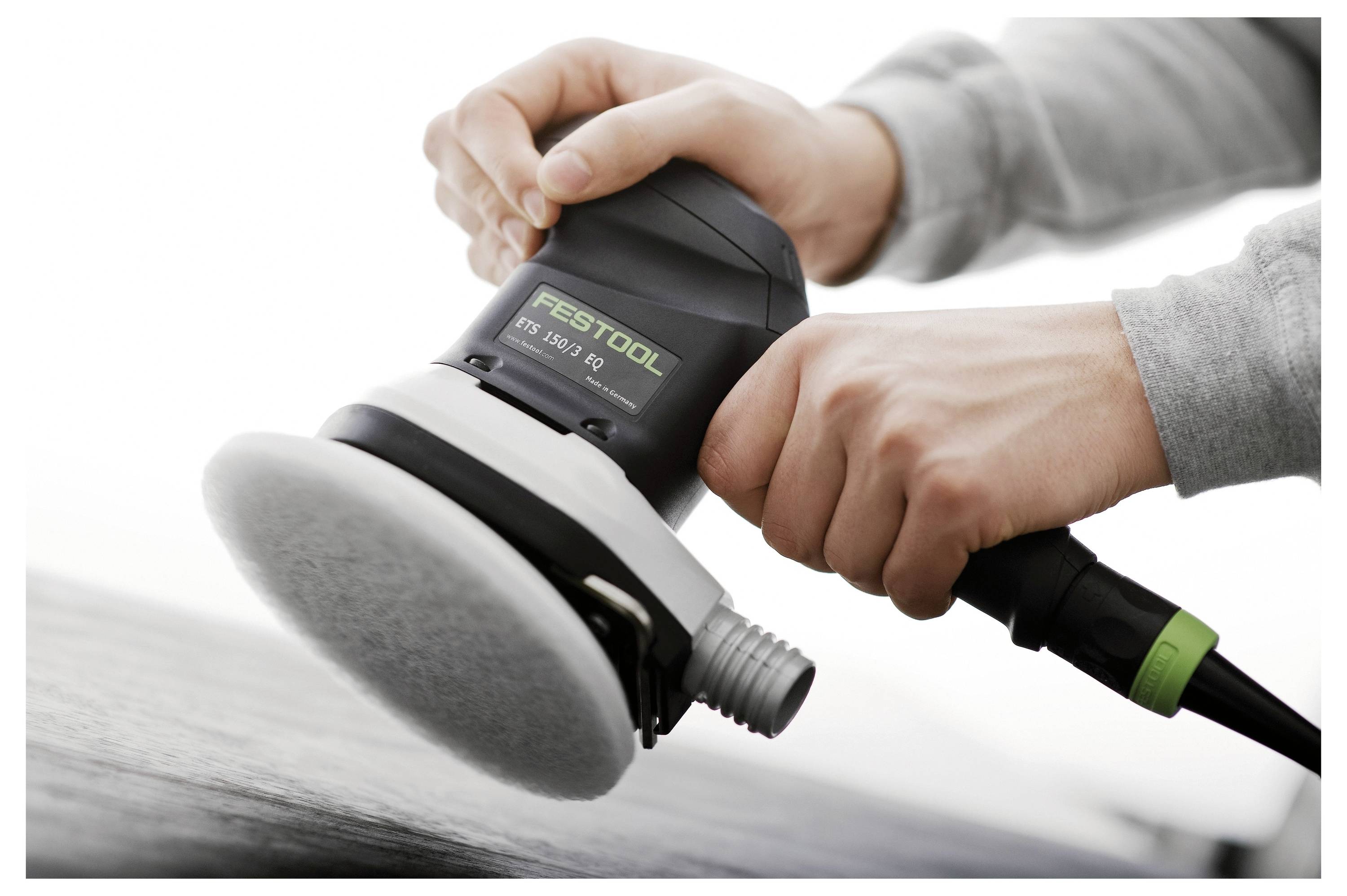 Két kéz egy 'Festool' márkájú elektromos csiszológépet tart, amely egy fa felületet simít. Az eszköz kábele látható, amely jobbra nyúlik.