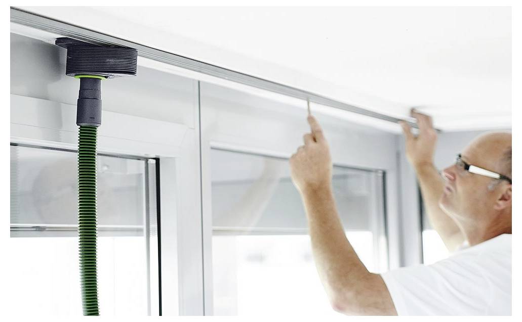 Egy személy mérőszalaggal méri meg az ablak keretét, precíz illeszkedésre koncentrálva. Az ablak fölött egy zöld tömlővel felszerelt szerszám van felszerelve.