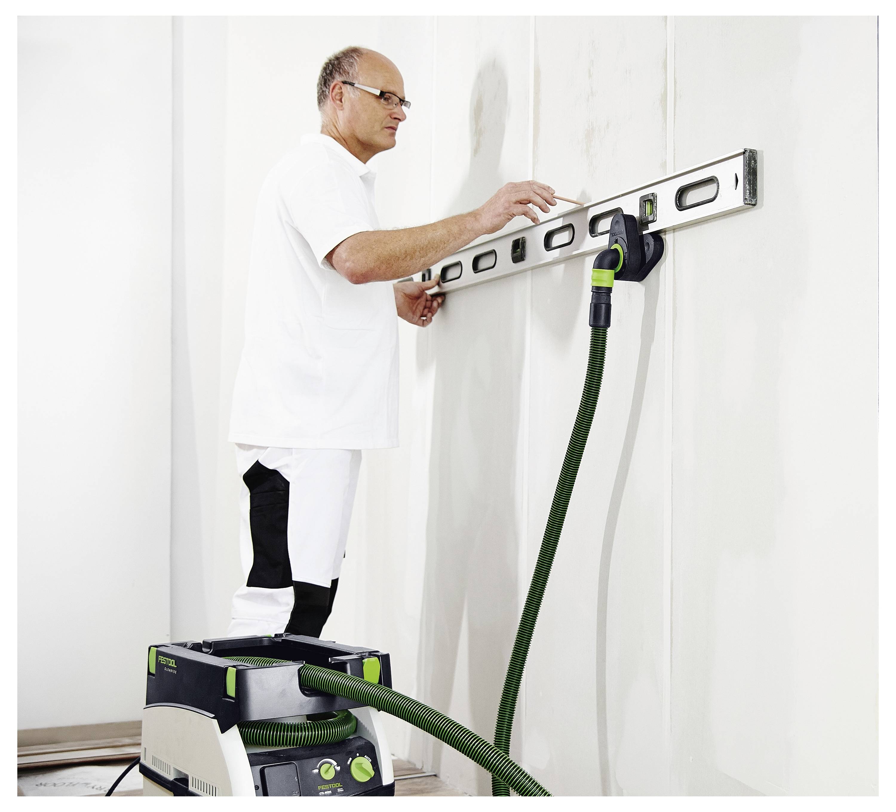 Munkás szintezőt használ egy fehér falon, egy lépcsős zsámolon állva; egy zöld csővel rendelkező porszívót használnak.