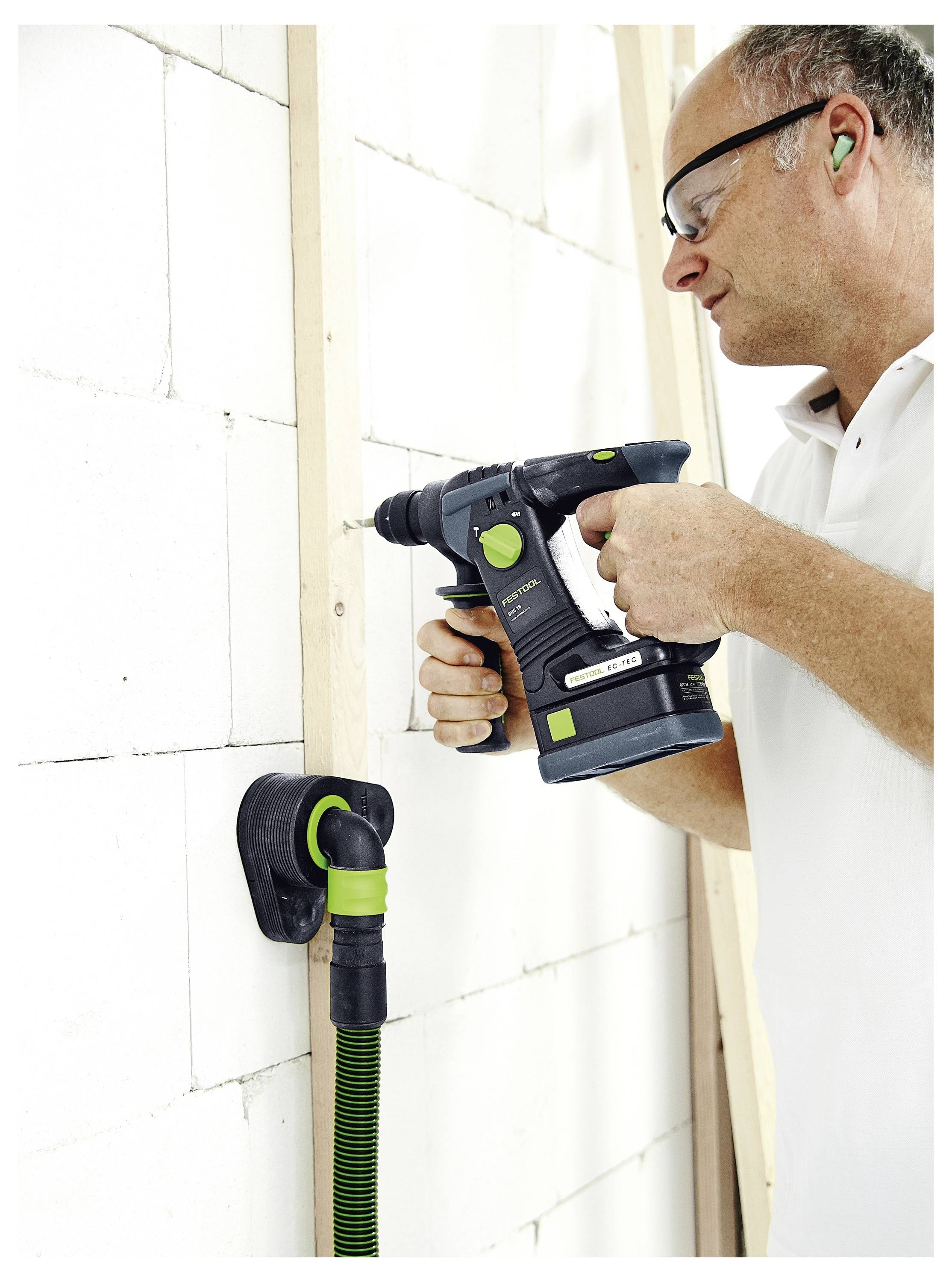 Egy személy vezeték nélküli fúróval egy fa deszkát rögzít egy fehér téglafallhoz, miközben biztonsági szemüveget és hallásvedőt visel.