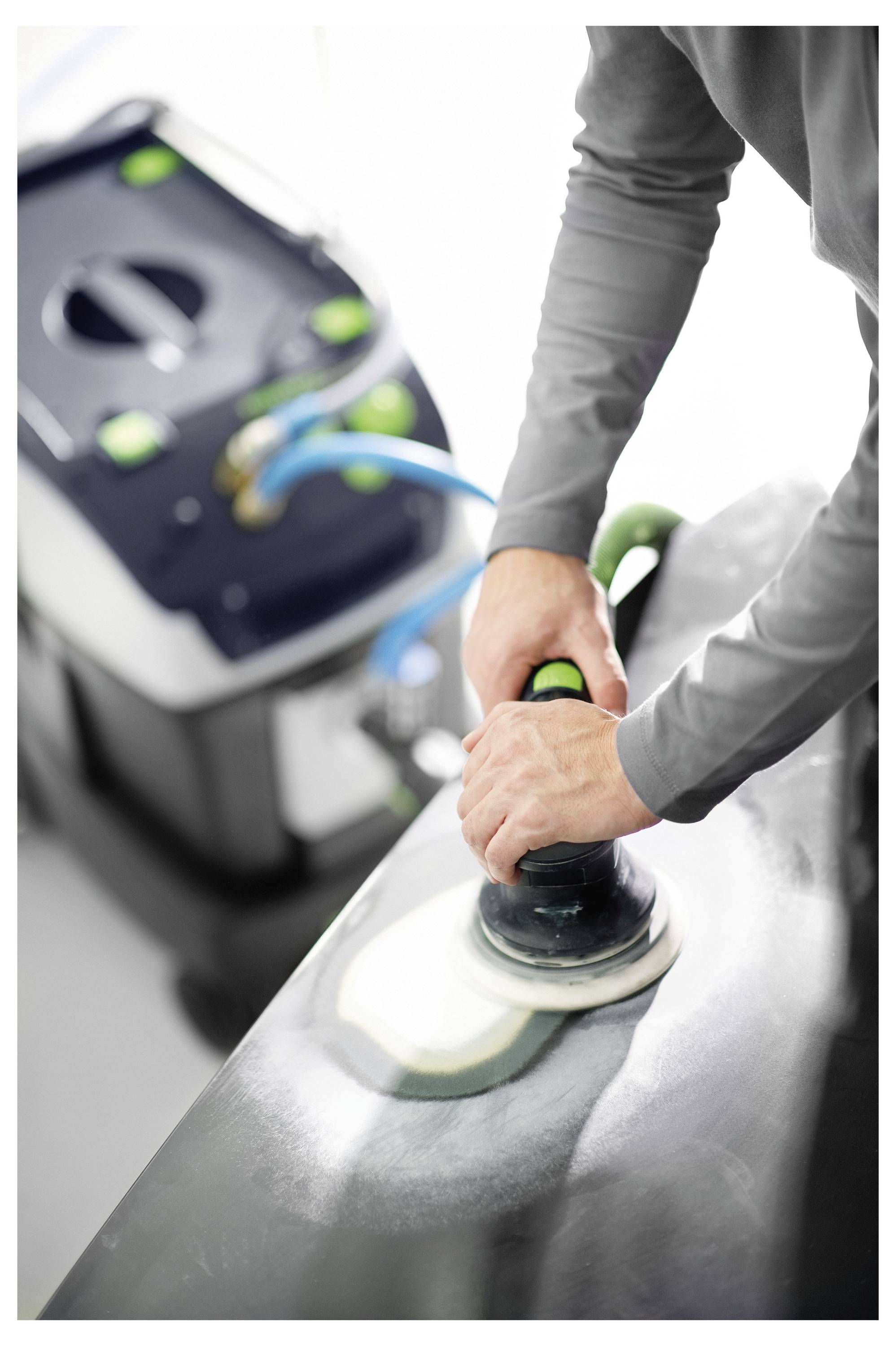 'Egy személy elektromos polírozógépet használ egy autó felületén, a háttérben pedig egy porszívógép látható, amely kék csővel csatlakozik.'