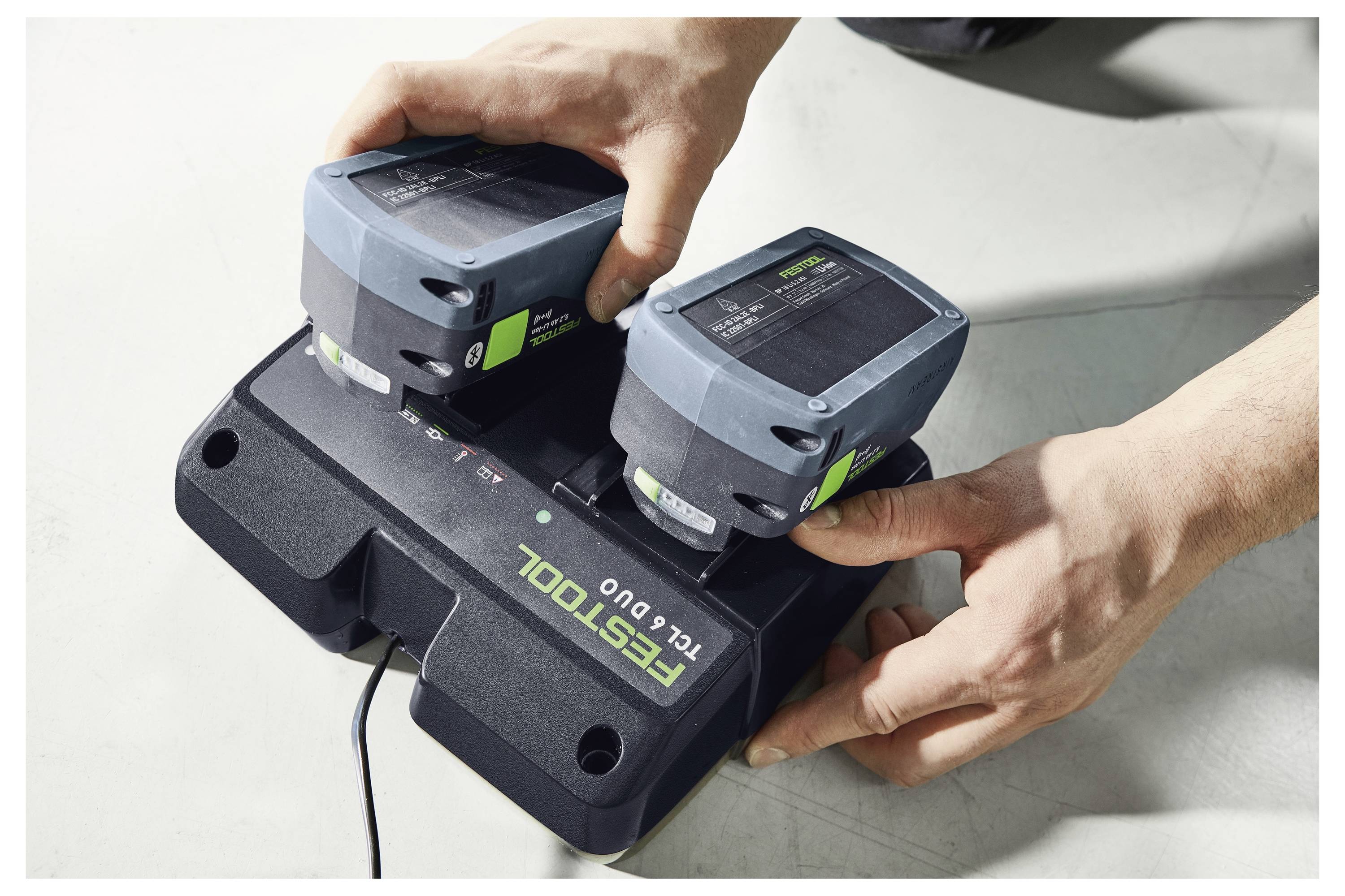 Két kéz két akkumulátort helyez el egy Festool TCL 6 Duo akkumulátor töltőre egy világos felületű felületre.