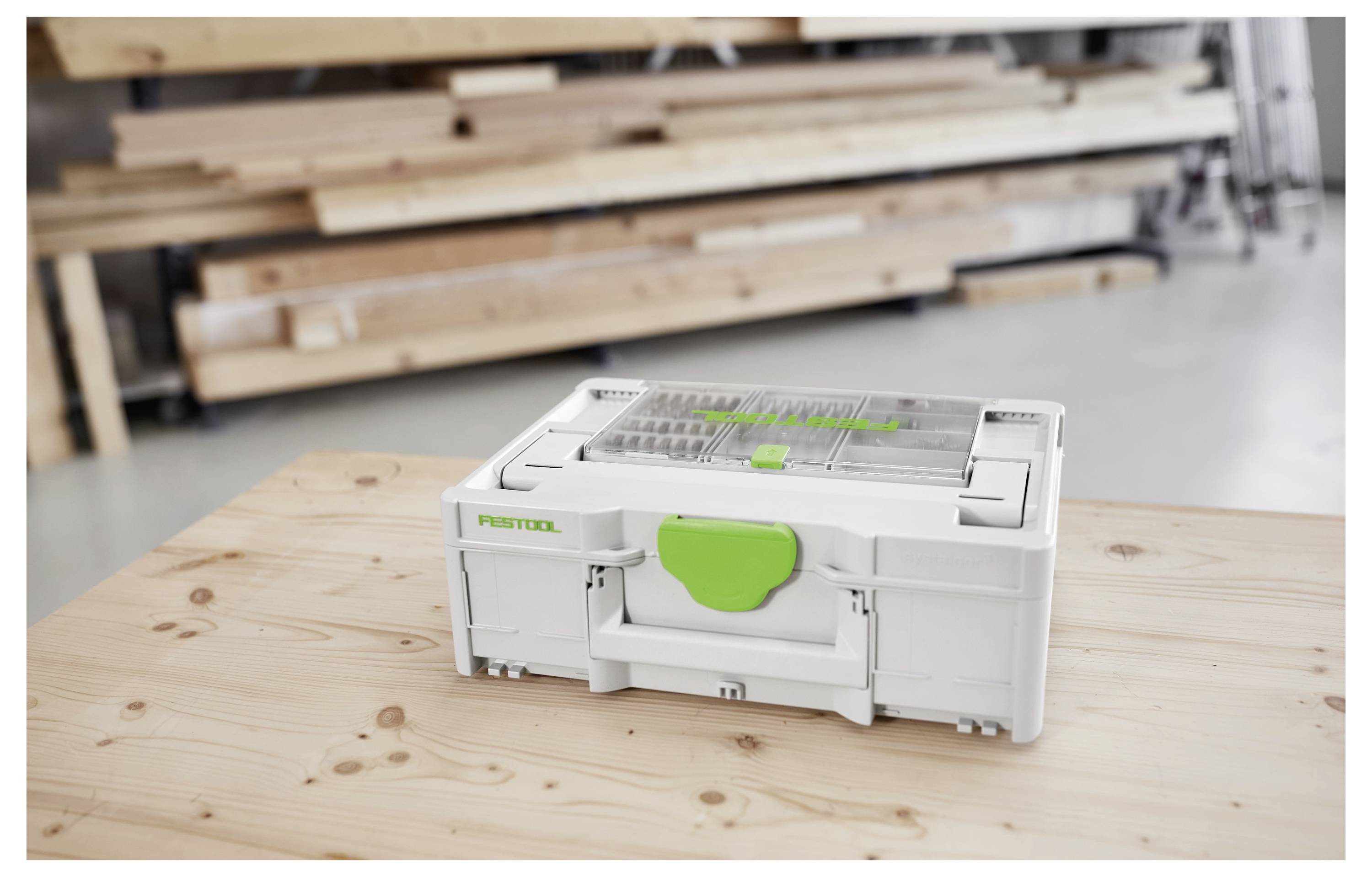 Egy kis, szürke tárológusztály, zöld retesszel és átlátszó fedéllel egy fából készült asztalon áll egy műhelyben, a háttérben elmosódott szerszámok és fádeszkák láthatók.