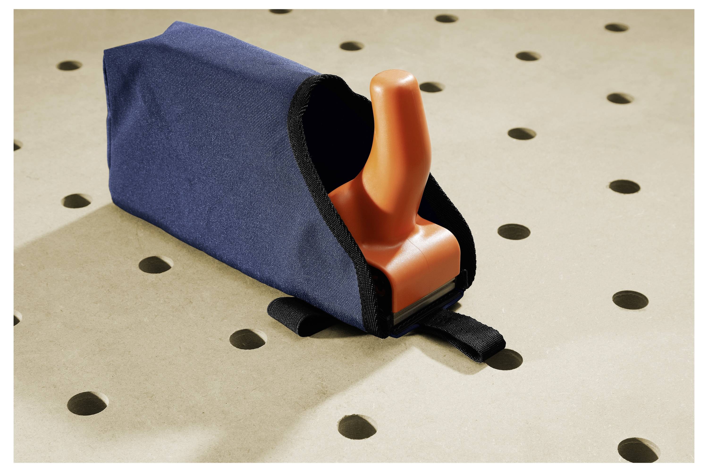 Narancssárga fogantyú, amely egy kék szövettel borított tárgyból nyúlik ki, amely egy perforált bézs felületen nyugszik.
