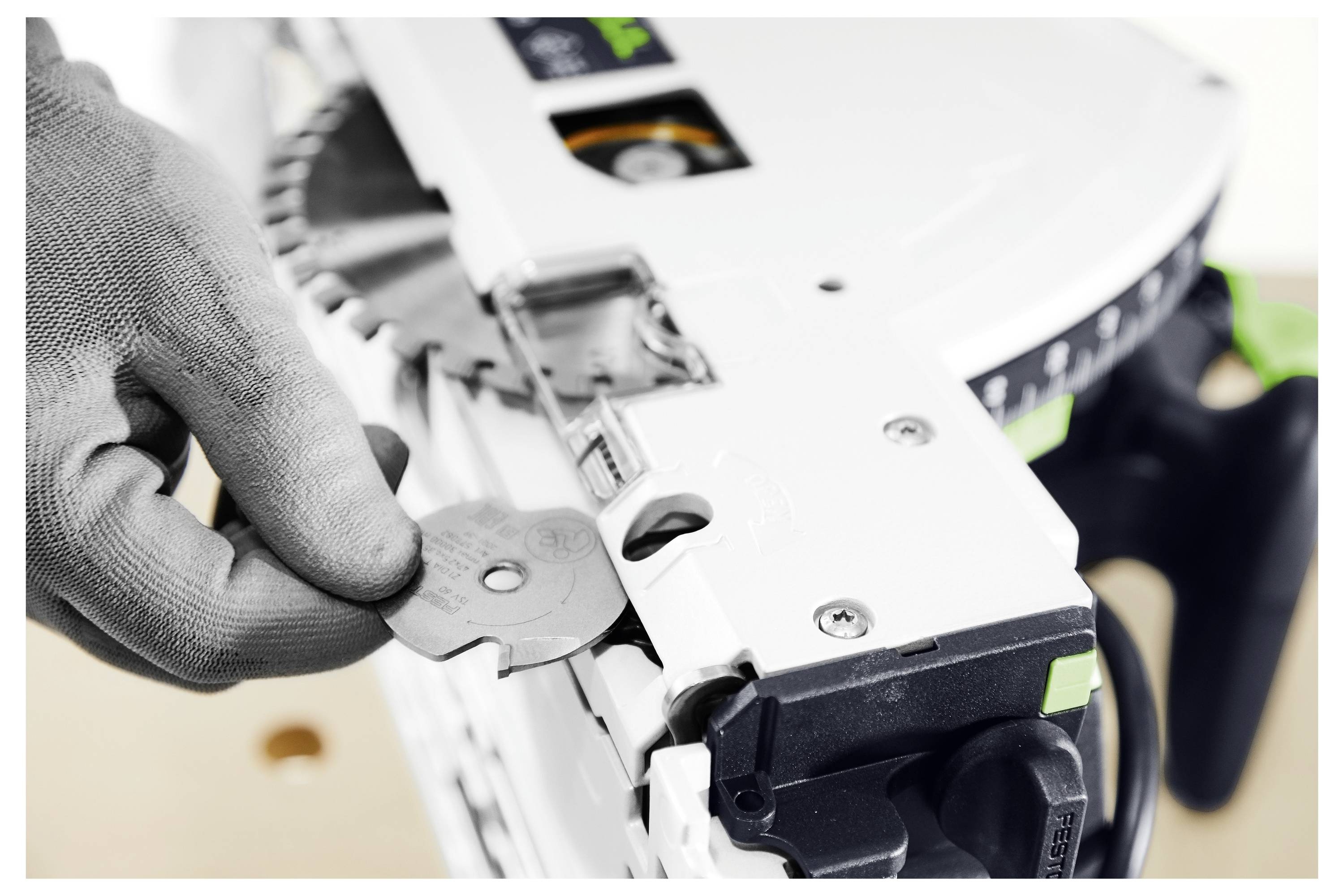 Egy kesztyűs kéz egy hatszögletű kulcsot helyez be egy fűrészbe a penge beállításához. A fűrész fehér, fekete és zöld kiegészítésekkel, a biztonsági funkciókra összpontosítva.