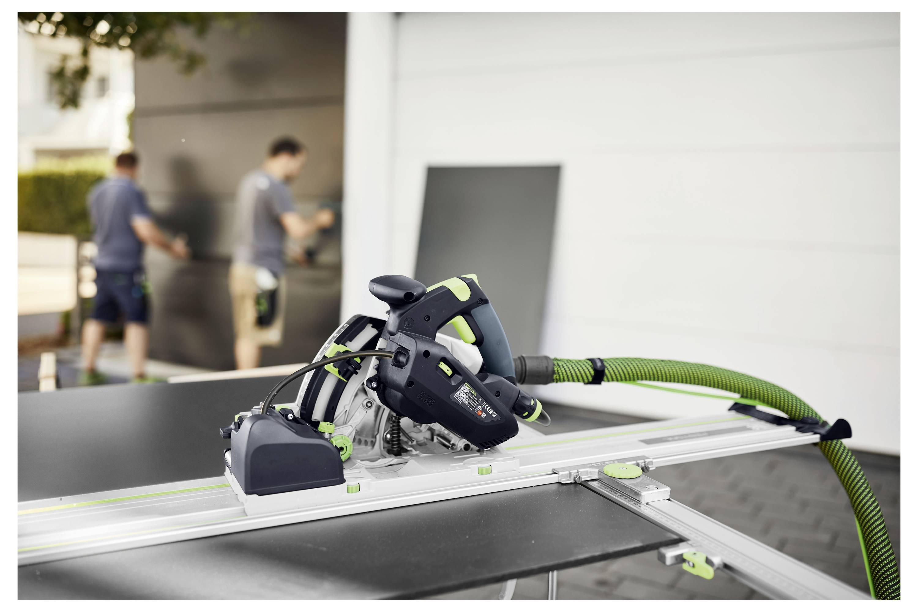 'Egy zöld tömlőcsatlakozással felszerelt körfűrész egy műhelyek asztalvágányán helyezkedik el. Két személy dolgozik a háttérben.'