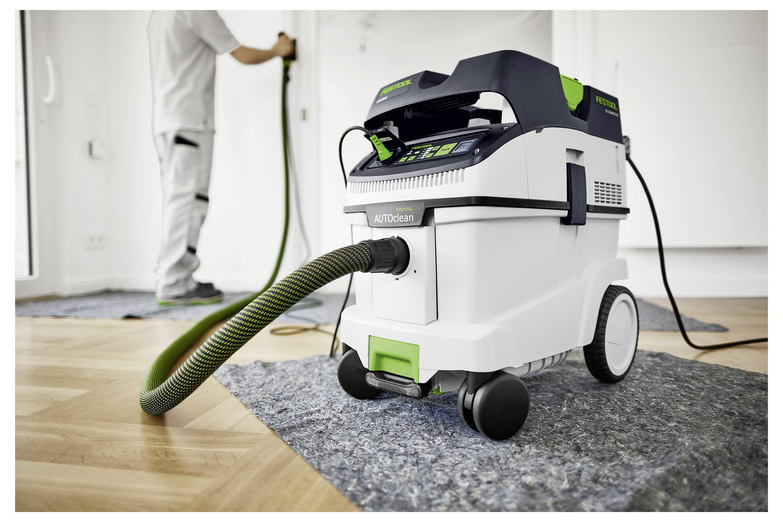 A porszívó csővel egy fa padlón van, és egy takarítási feladatra összpontosít. A háttérben egy személy használja a porszívót.
