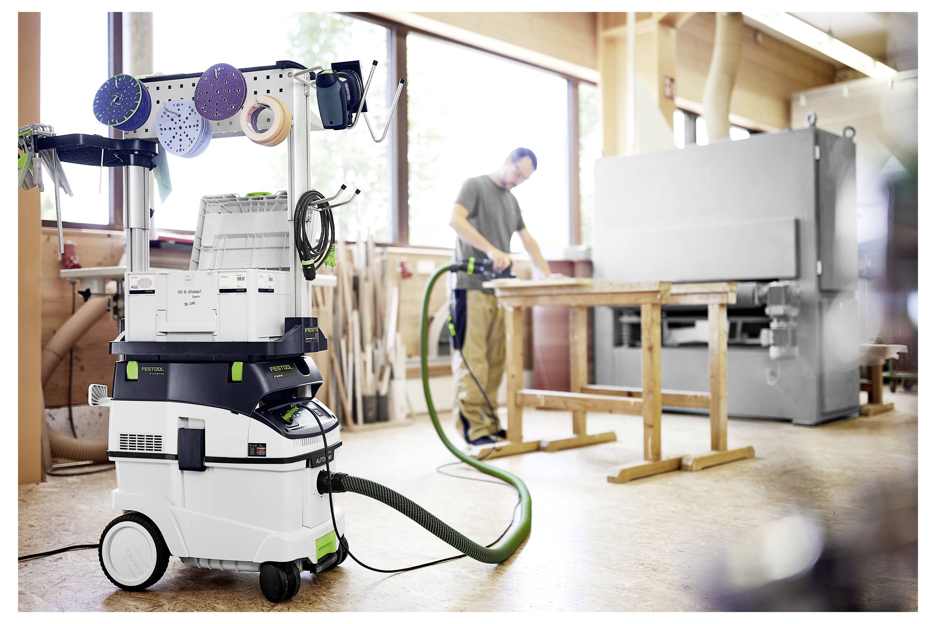 Egy asztalos porcsiszolóépítést használ egy műhelyben; egy porgyűjtő áll az előtérben, amely egy csővel csatlakozik a géphez.