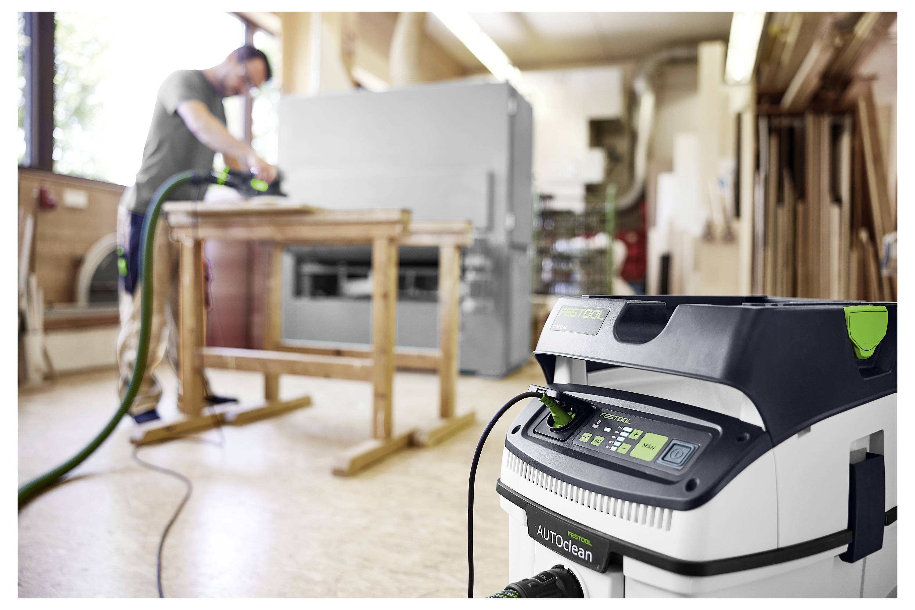 „AUTOclean" porszívó előtérben; személy fa csiszolásával foglalkozik a műhelyben a háttérben, amely a hatékony porgyűjtésre való fókuszt sugallja.