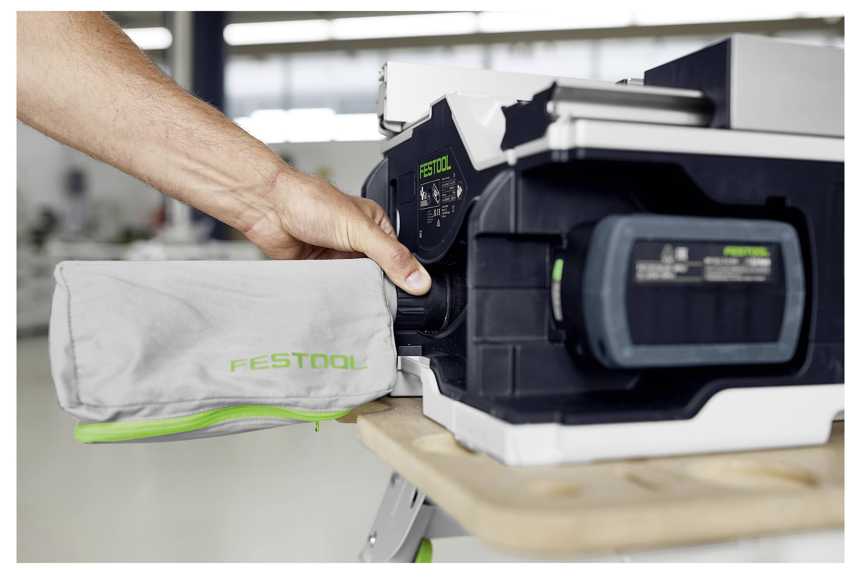 Egy személy egy porzsákot erősít rá egy Festool elektromos szerszámra egy műhelypulton a műhelyben.