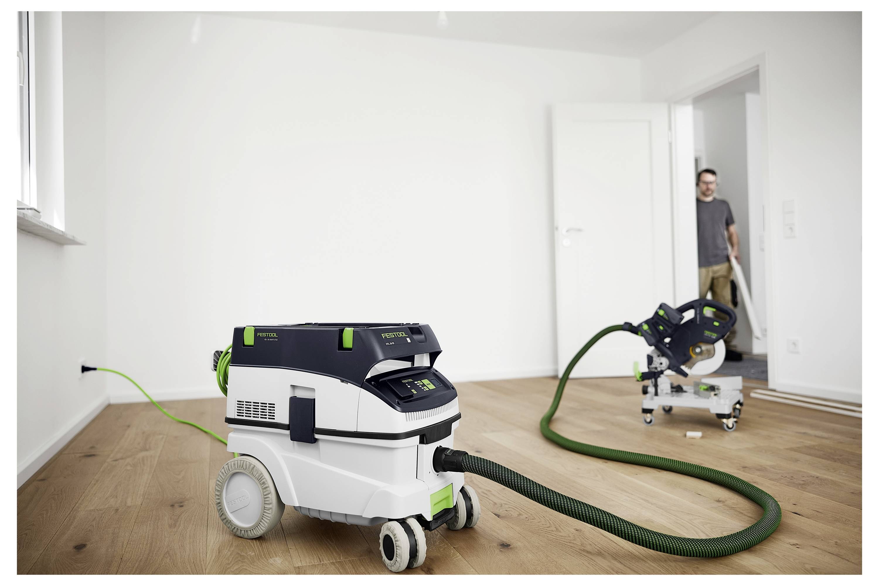 Egy nagy kerekeken lévő porszívó zöld csővel az előtérben fa padlón áll. Egy személy áll egy ajtónyílásban a háttérben.