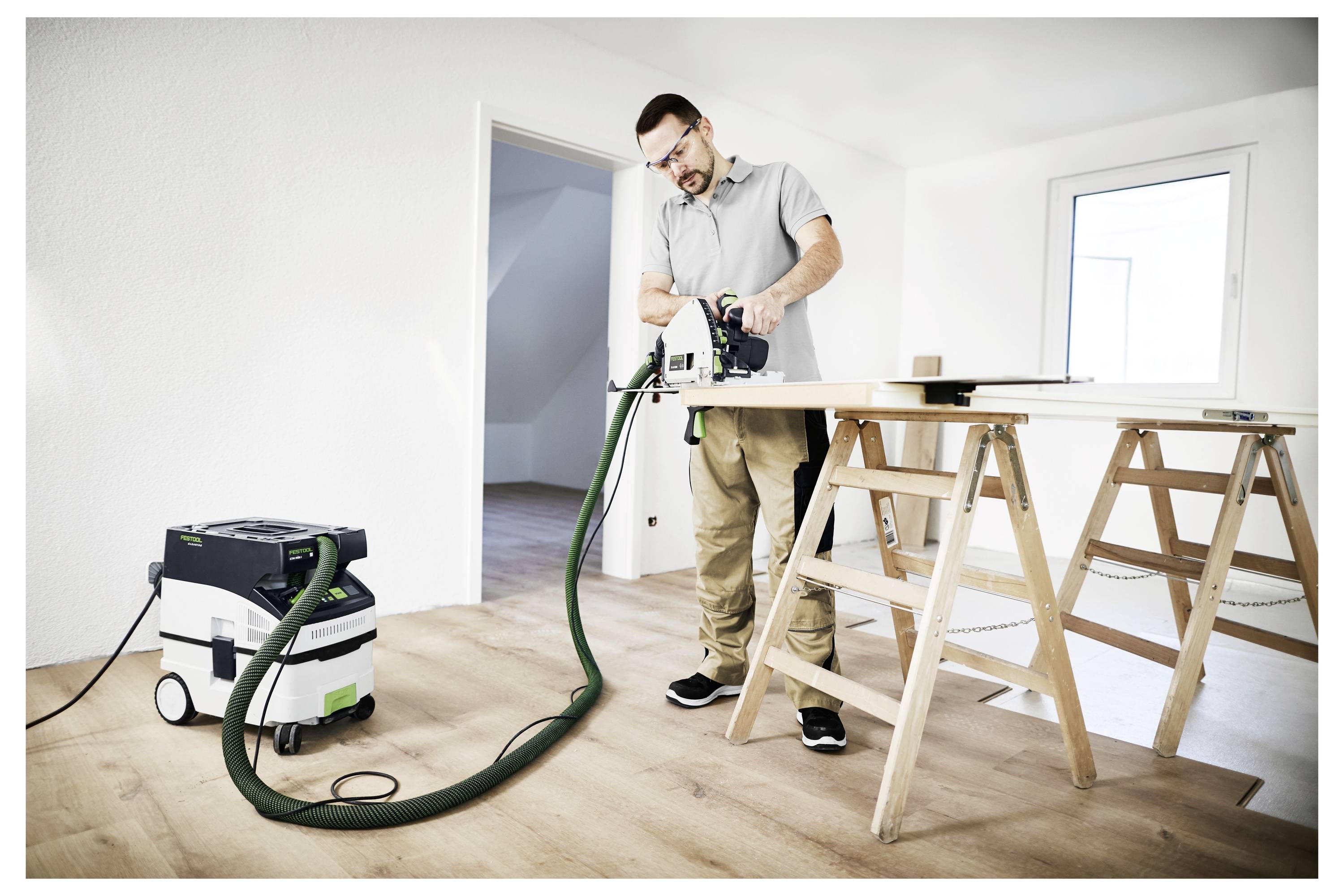 Egy biztonsági szemüveget viselő férfi elektromos fűrésszel dolgozik egy fa deszkán, amelyet szalmakosarak támasztanak alá egy jól megvilágított, fehér falú szobában.