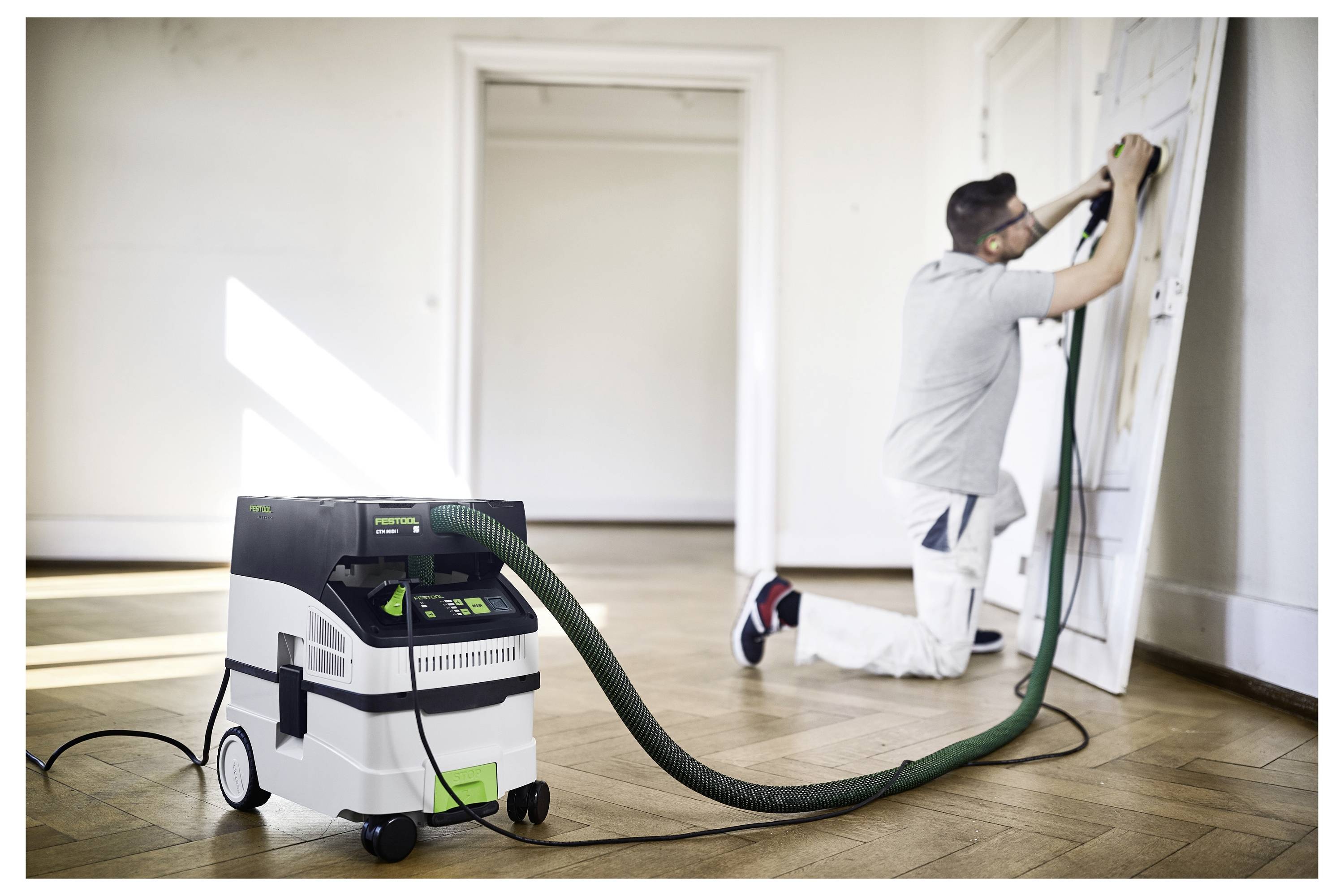 Egy munkaruházatban lévő személy egy ajtót csiszol elektromos szerszámmal, amely egy fehér és zöld porgyűjtőhöz csatlakozik, egy világos szobában fa padlózattal.