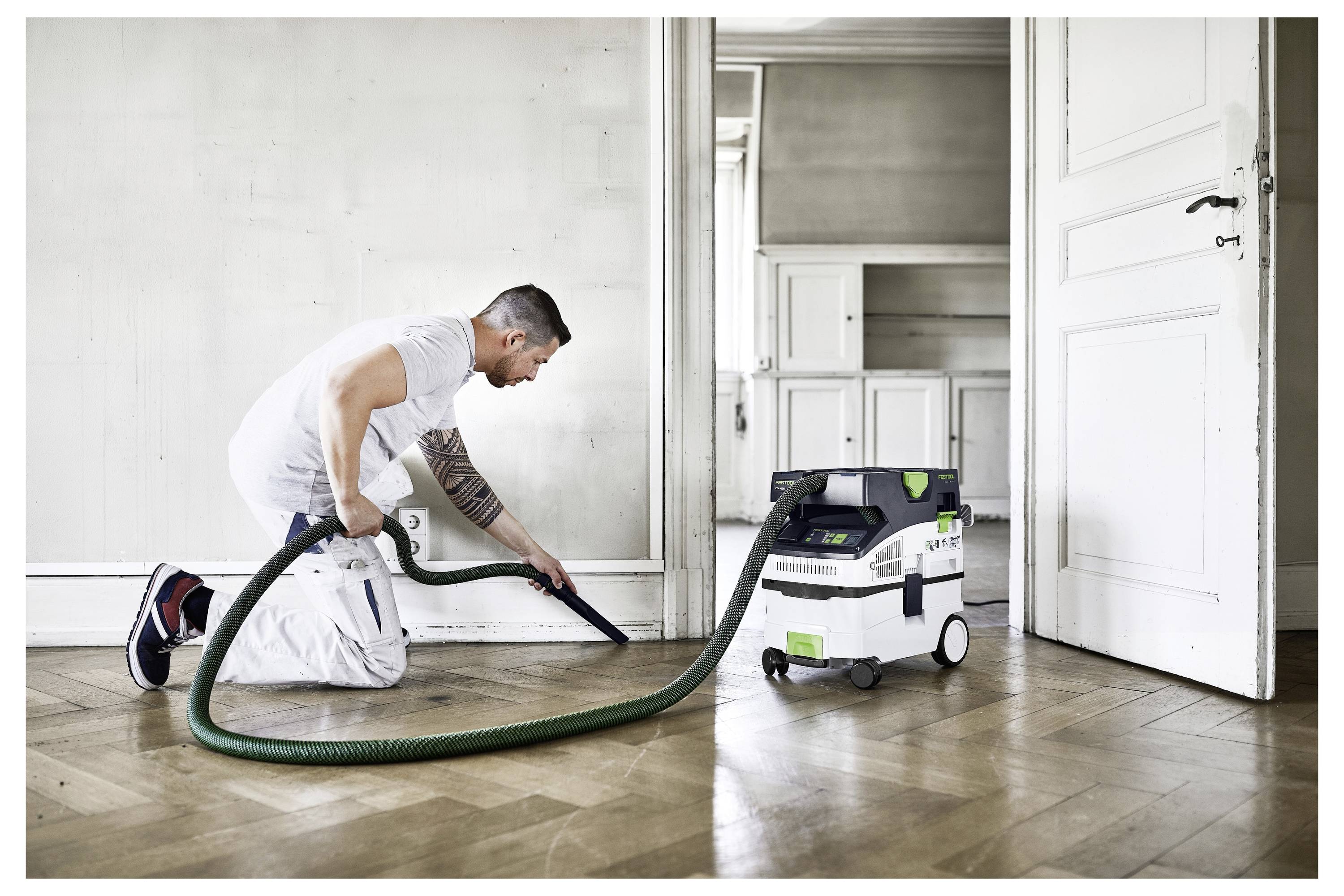 Egy személy porszívót használ egy fa padlóval rendelkező szobában fehér falakkal és nyitott ajtókkal, amely takarítást vagy otthoni karbantartást sugall.