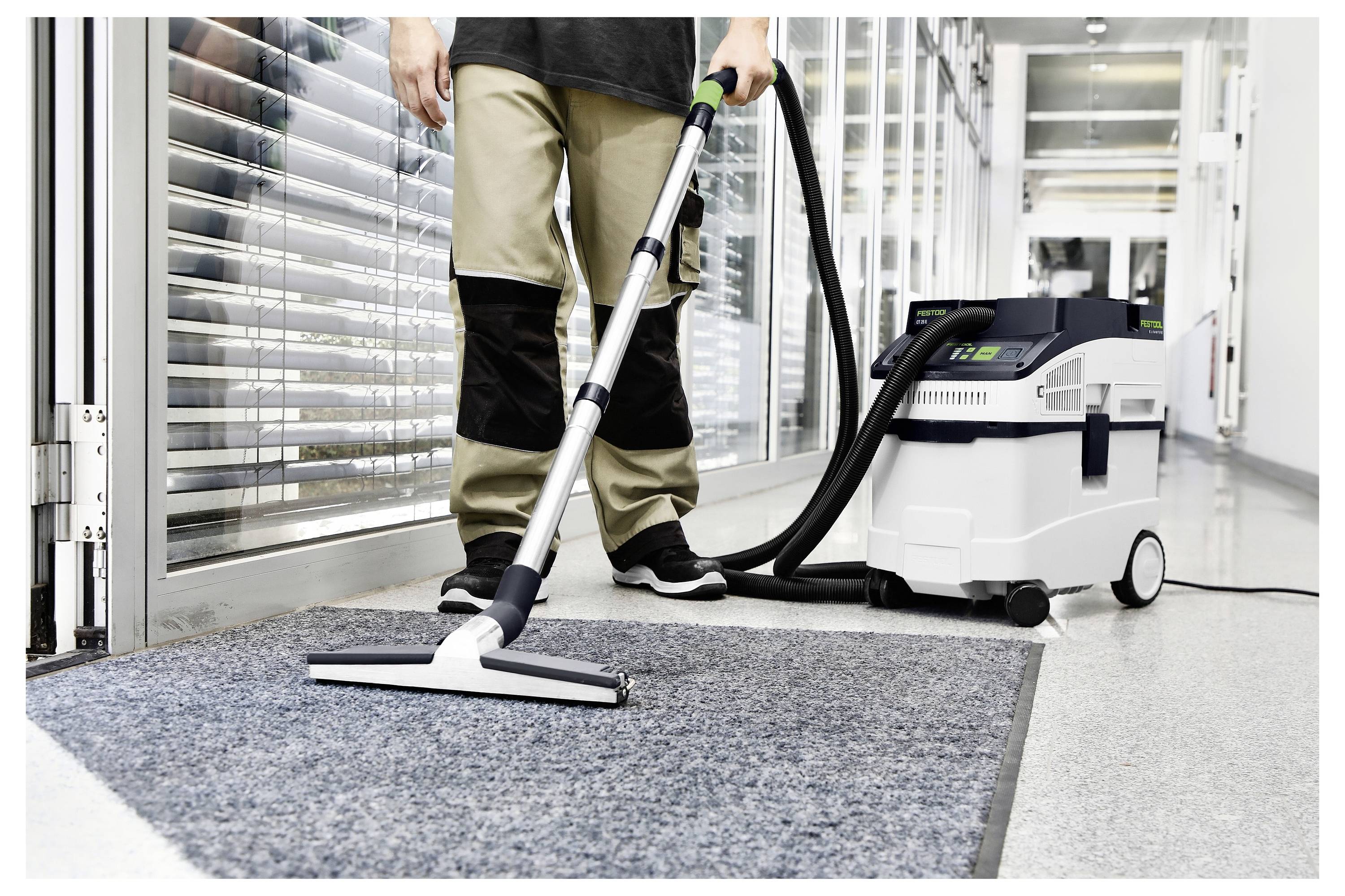 Egy személy egy hosszú folyosó szőnyegét takarítja meg egy ipari porszívóval, fekete és barna munkanadrágban és fekete cipőben, nagy ablakok közelében.