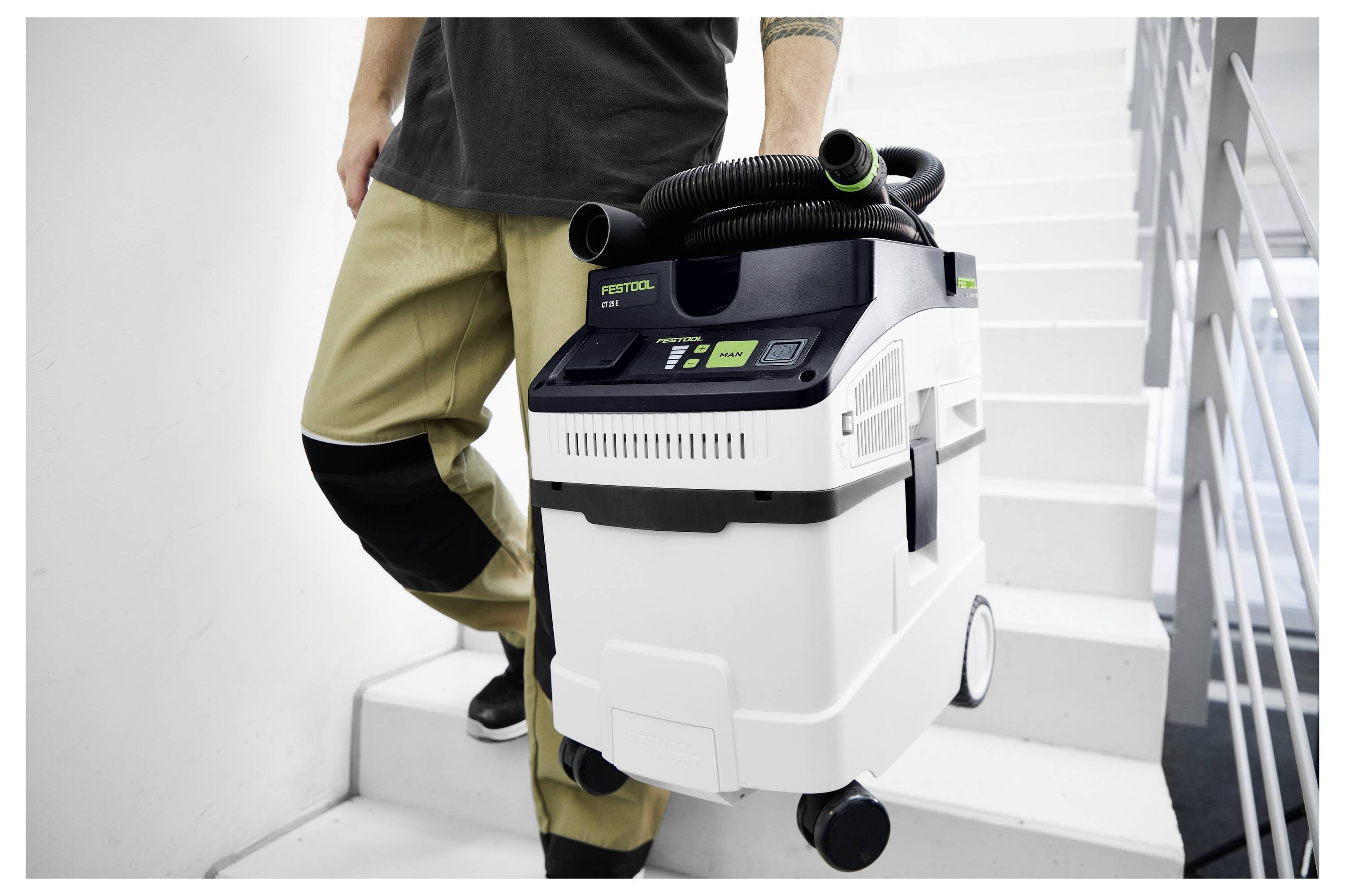 Egy szürke póló ingben öltözött személy áll egy faipari anyagokkal teli műhelyben, egy elektromos szerszámot tart a kezében, és mosolyogva néz a kamerába.