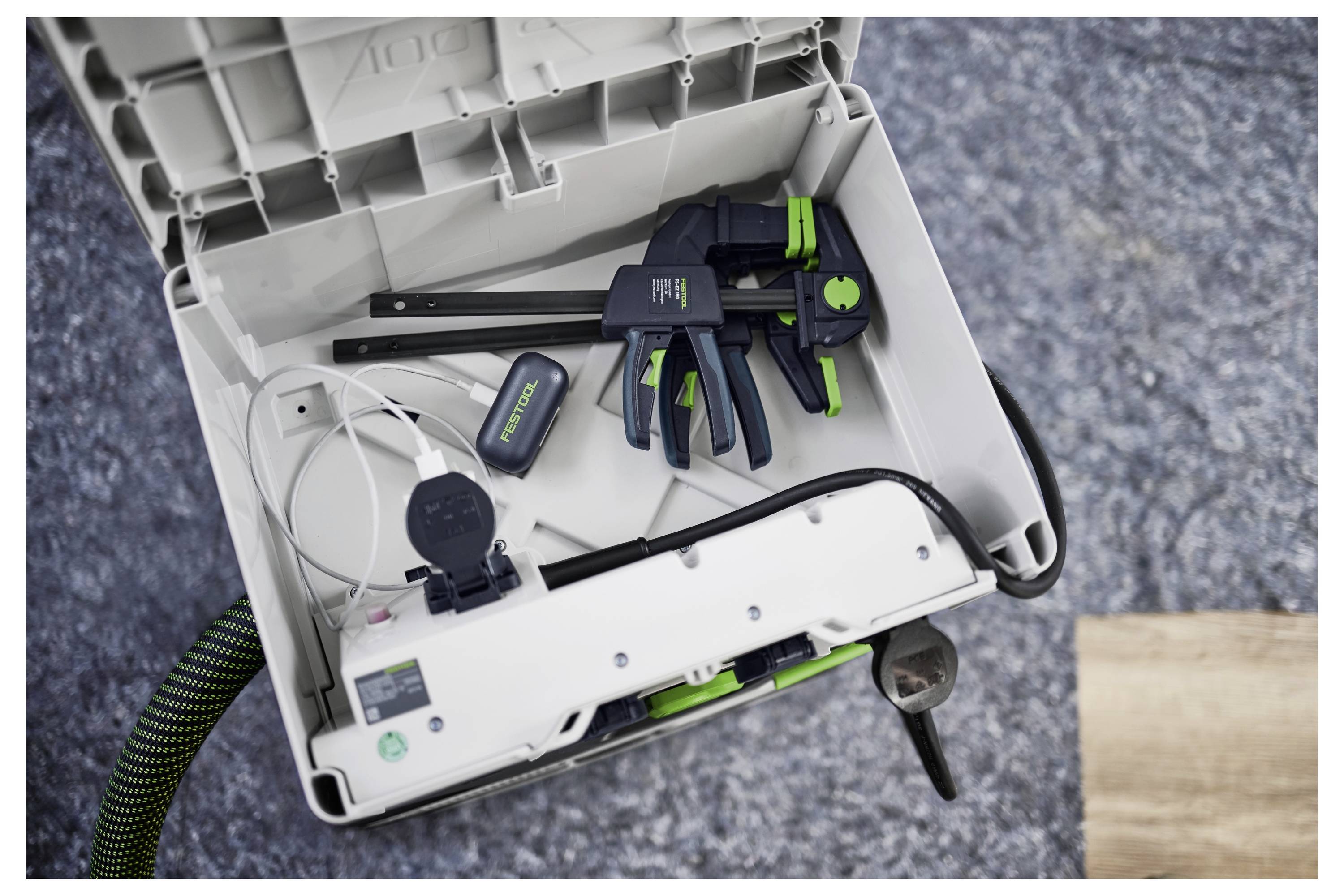 Nyitott szerszámosdoboz fogókat, egy digitális kijelzőegységet és kábelekat tartalmaz egy szürke felületen. A háttérben táblák és műhelyi szerszámok láthatók.