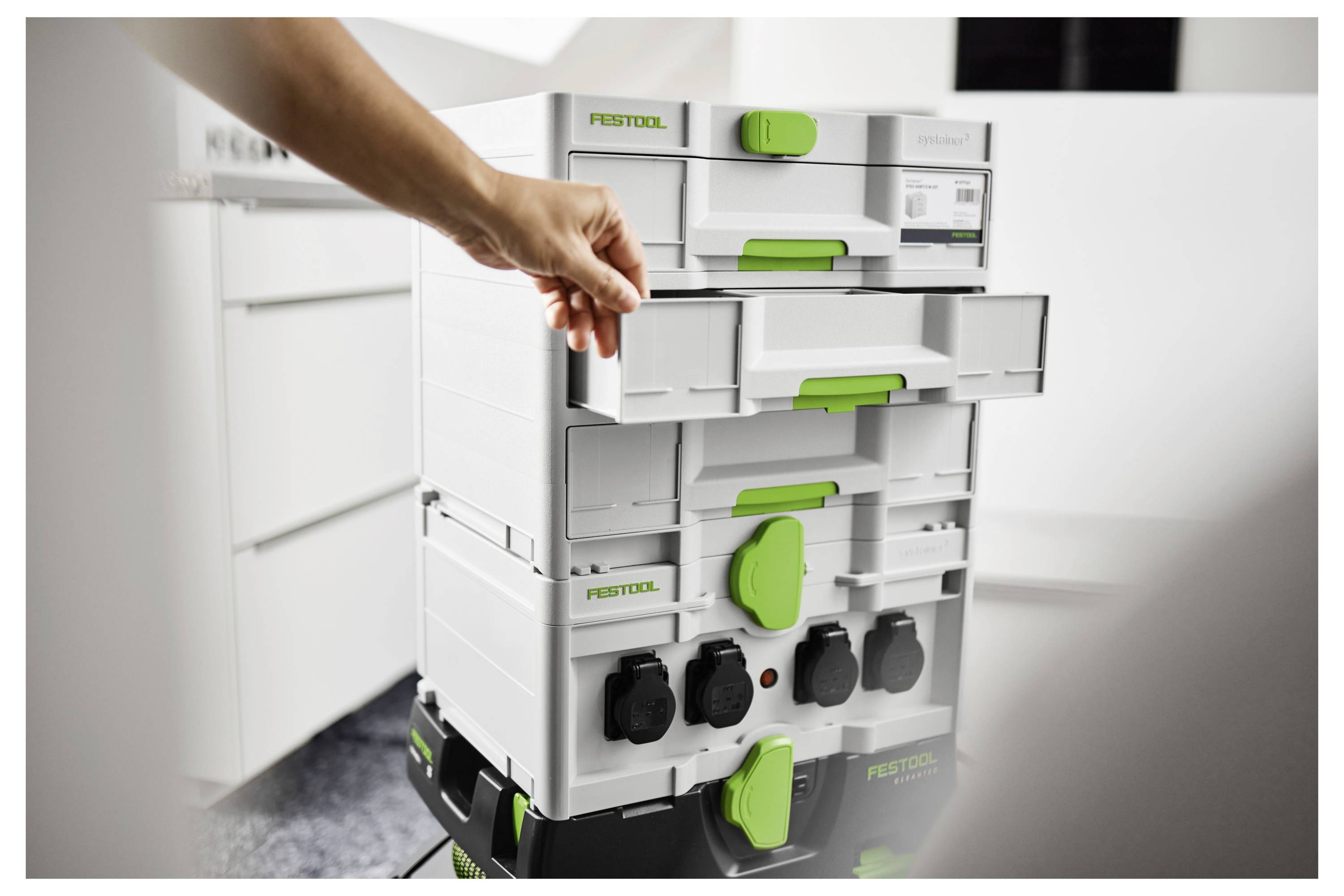 Egy kéz egy fiók-t nyit meg egy fehér és zöld Festool tárolóegység-készlet-en egy műhelyben, amely szervezett szerszámtárolást mutat.