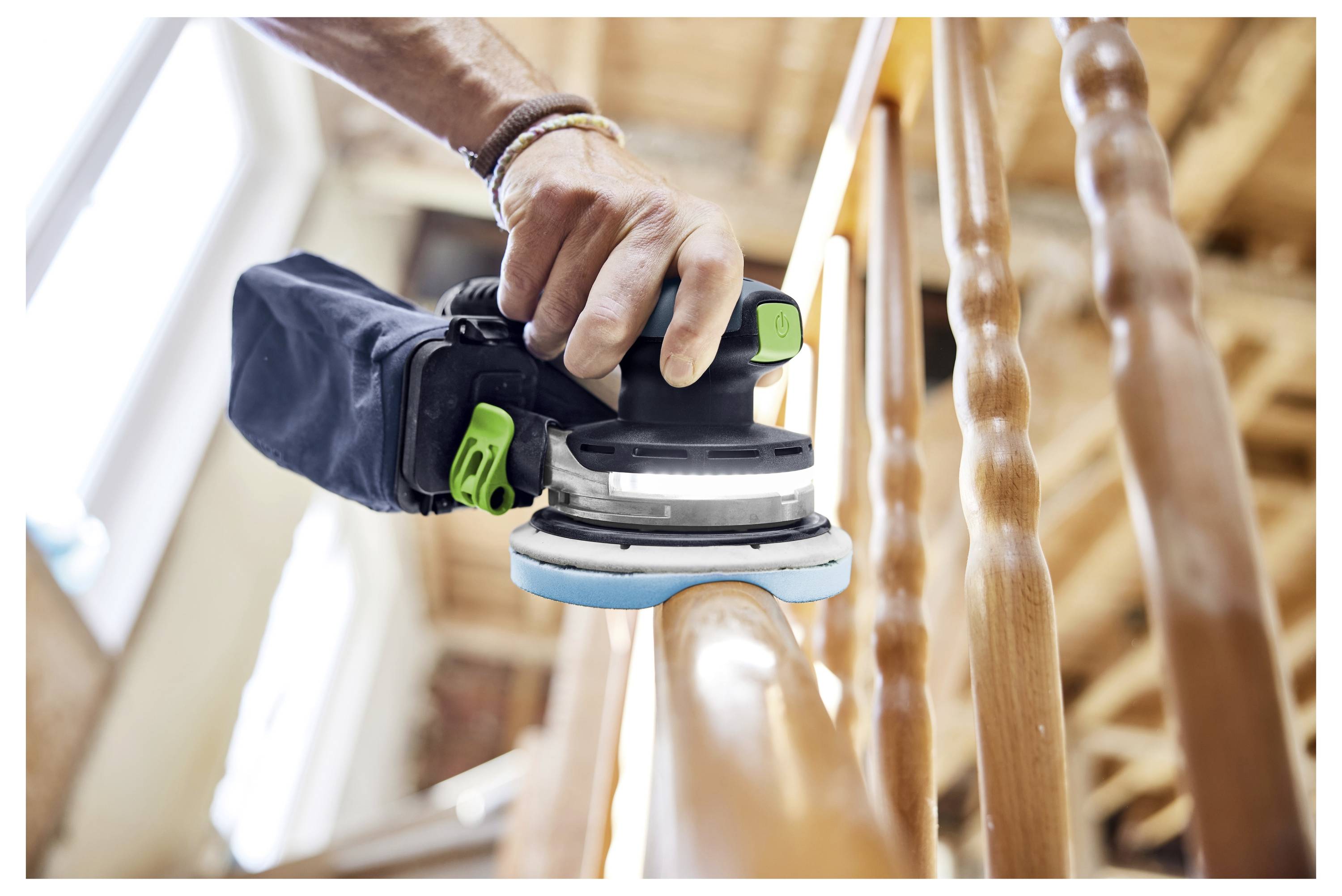 Egy személy kézzel tartott elektromos csiszolóval egy fa lépcsőkorlátot simít egy szobában, ahol fa gerendák vannak.