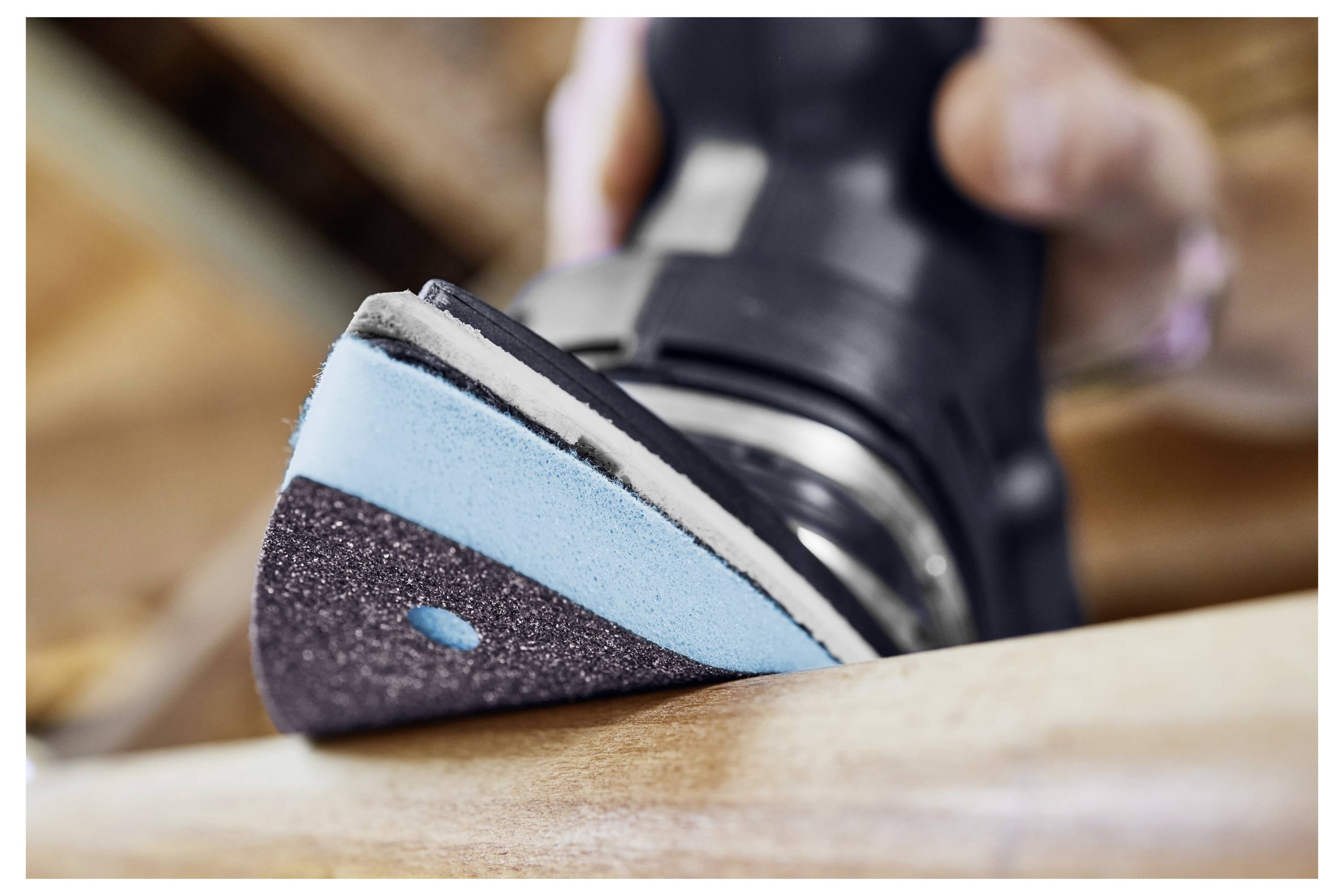 Egy közeli képet mutat egy kéz, amely egy elektromos csiszológépet tart, és egy fa deszka felületét simítja, feltárva a kék és fekete csiszolókorongot.