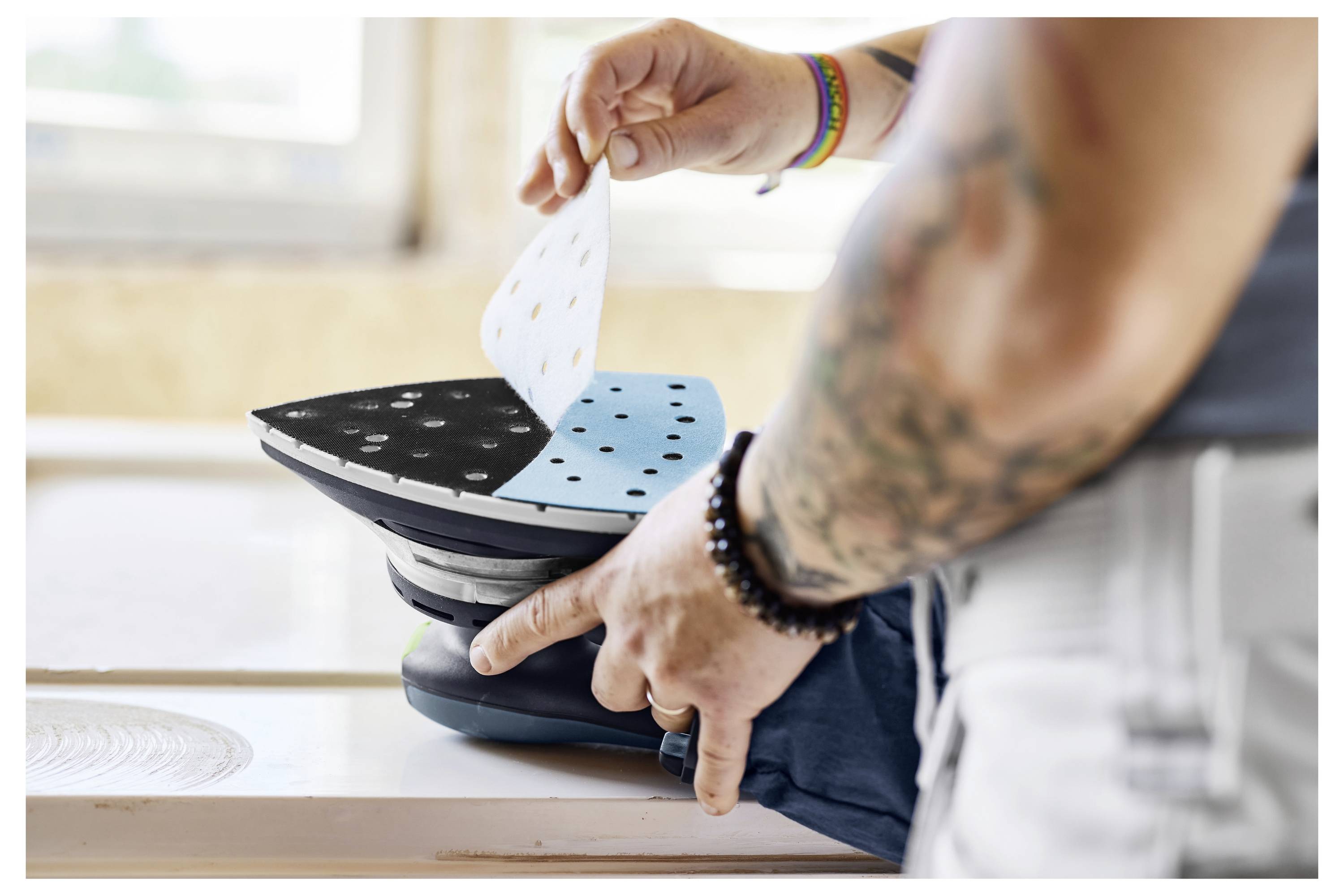 Egy személy csiszolópapírt helyez egy csiszolószerszámra, előkészítve azt a használatra. A személy karján látható fényes csuklópántok és tetoválások.