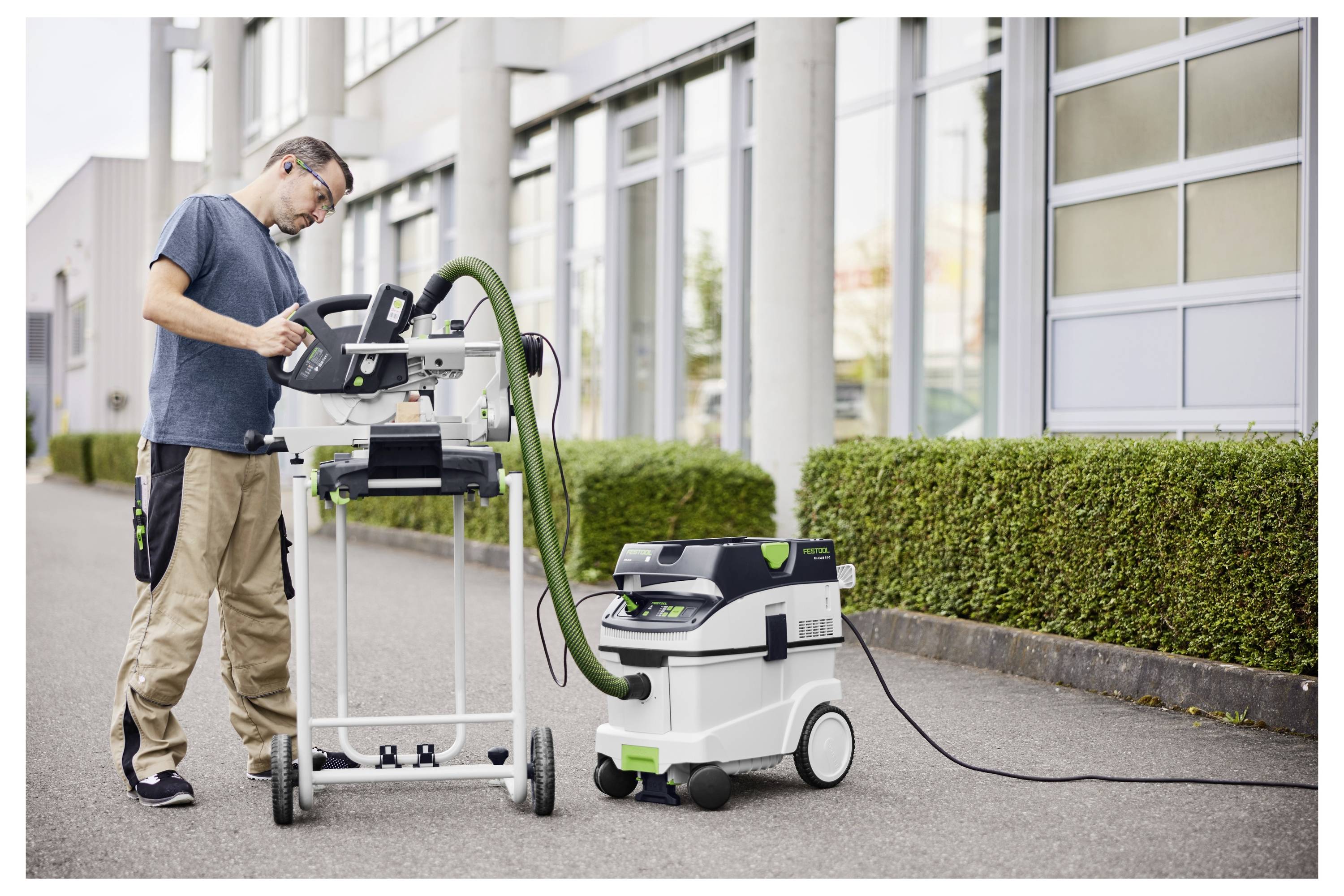 Egy személy asztali fűrésszel vág fát egy műhelyen belül. Védőszemüveget és szürke kapucnis pulóvert visel, és teljes odafigyeléssel végezte a feladatát.