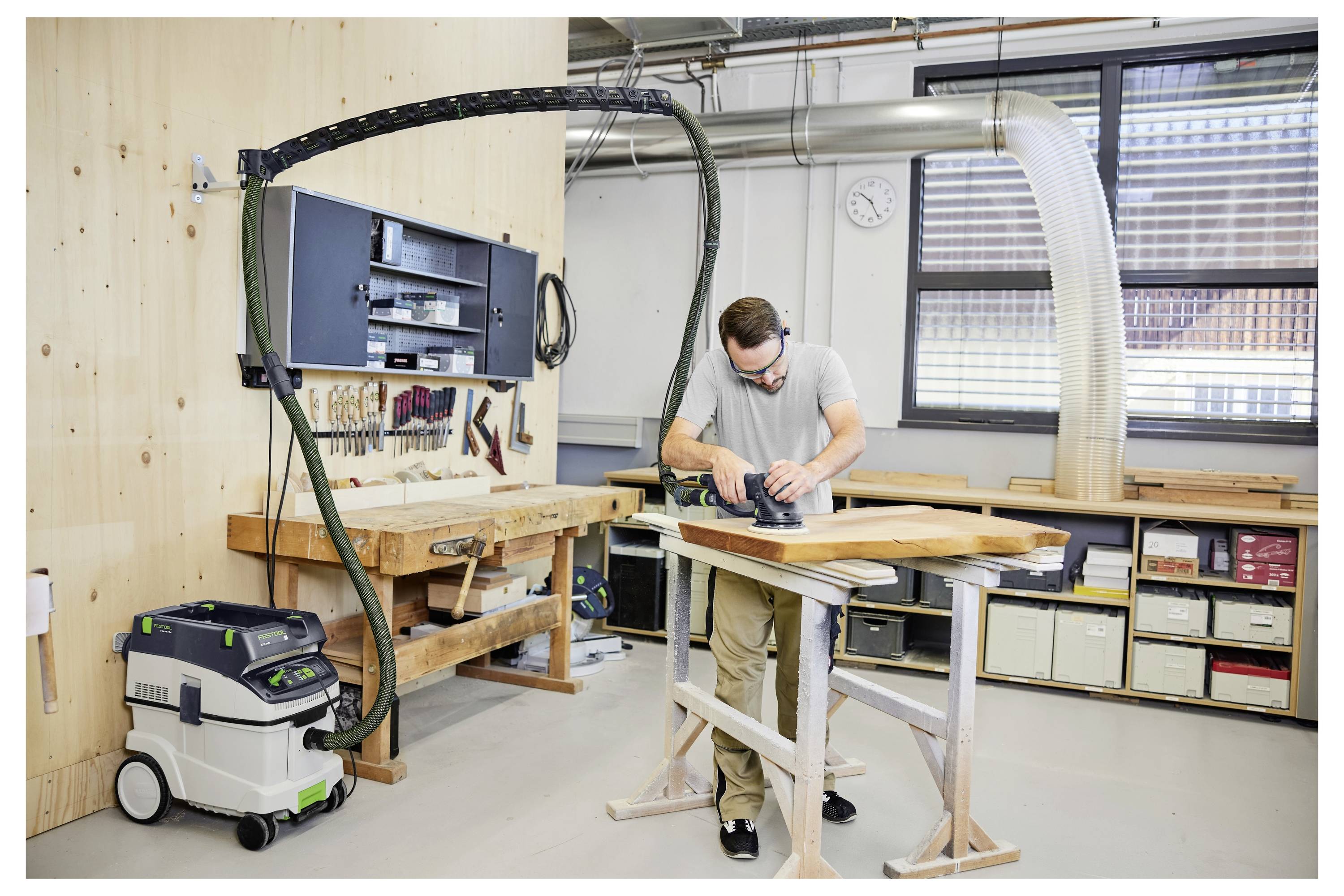 Egy személy elektromos csiszolót használ egy fadeszkán egy olyan műhelyben, amely szerszámokkal, munkapadokkal és egy porgyűjtő rendszerrel van felszerelve.
