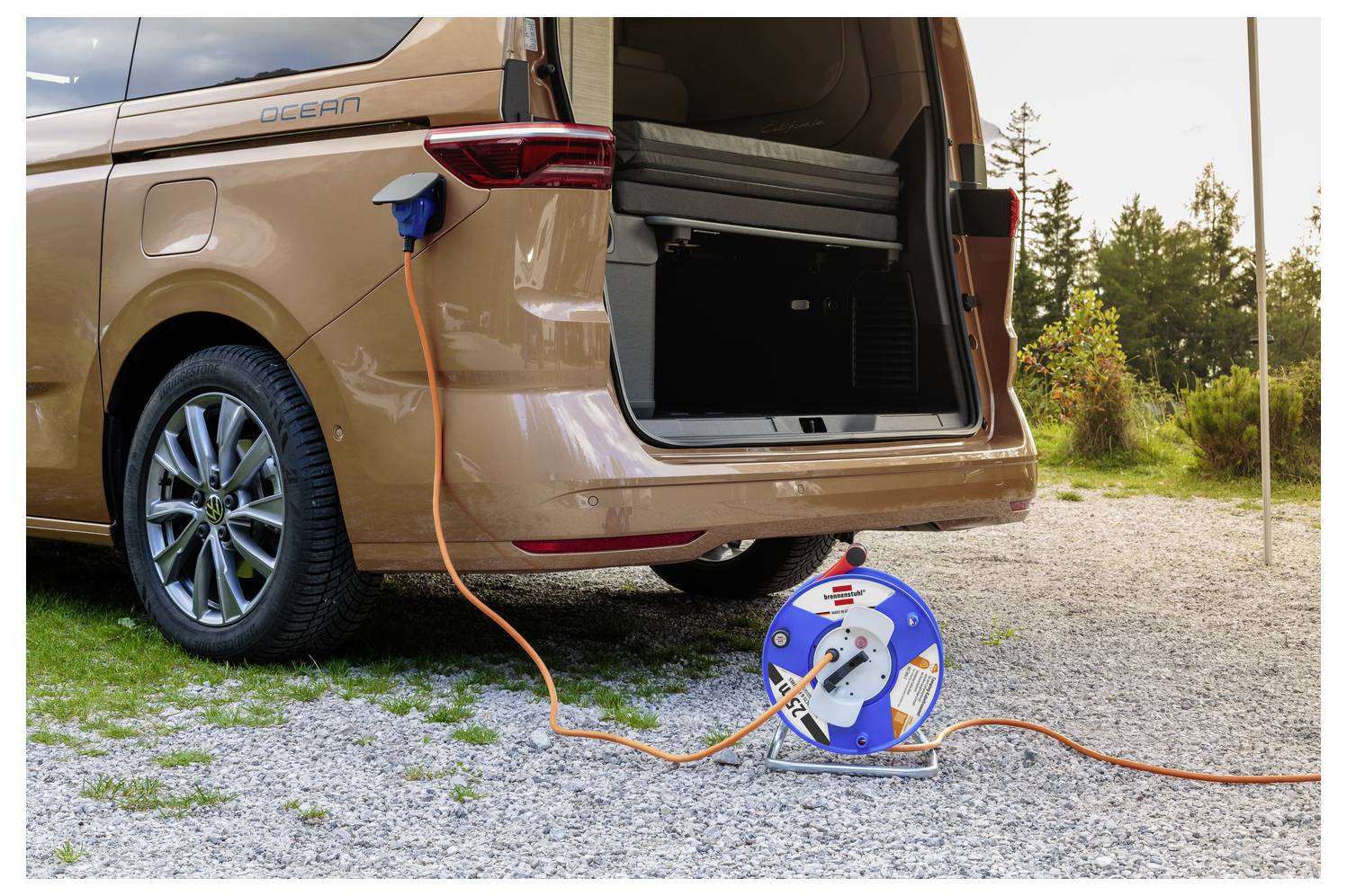 Elektromos kisbusz egy töltőkábelbe dugva kívül, egy hordozható kábeltekercsel a nyitott hátsó ajtó mellett a kavicspadon.