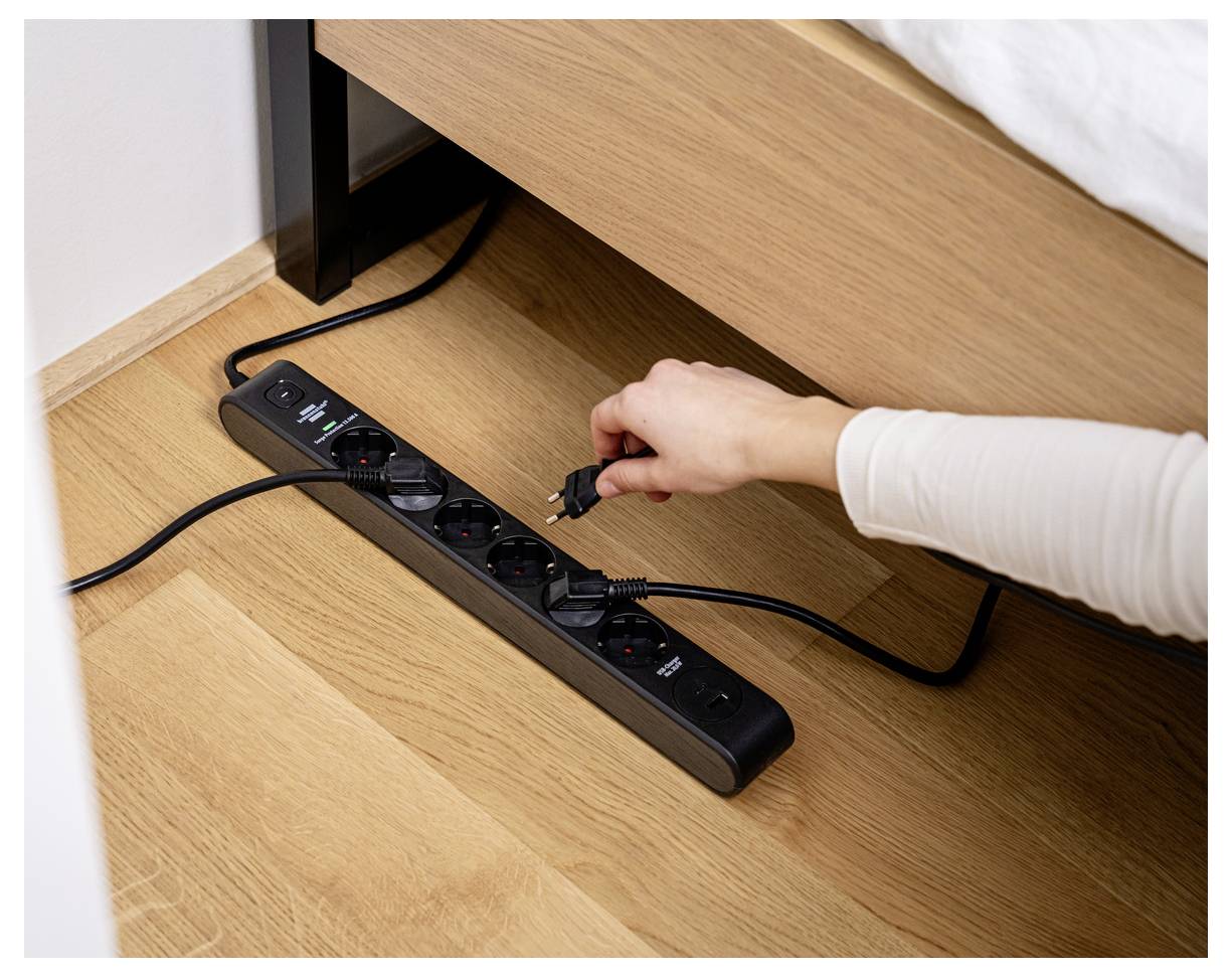 Egy kéz egy kábelt dugmál be egy fekete szalag talapzatba egy fapadlón, amely egy ágy mellett található, az elektromosság felhasználására és a kapcsolatra összpontosítva.