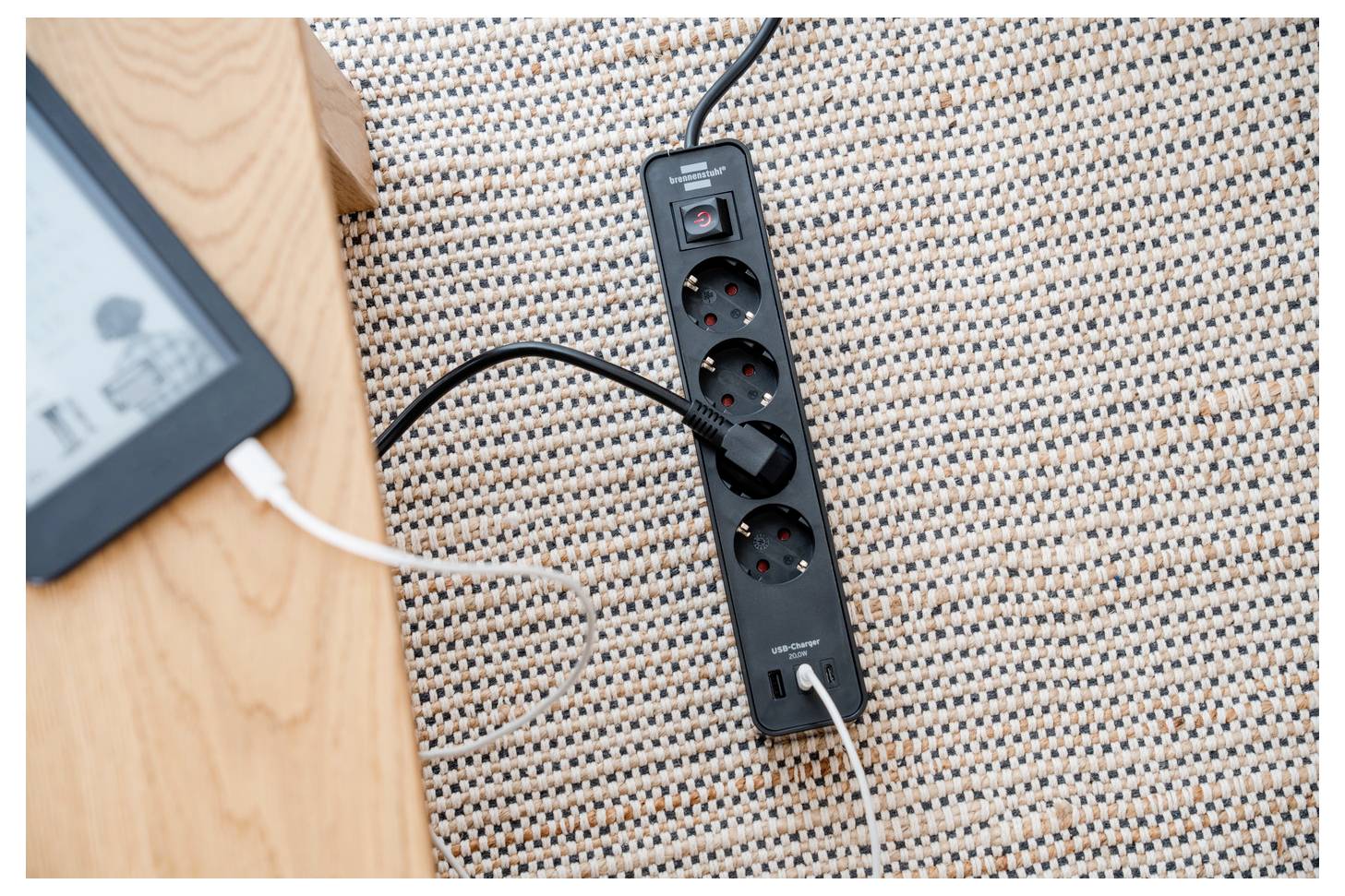 Elosztósor egy szőtt szőnyegen egy fa asztal mellett, több bedugaszolt kábellel, ami elektronikus eszközök töltésére utal.